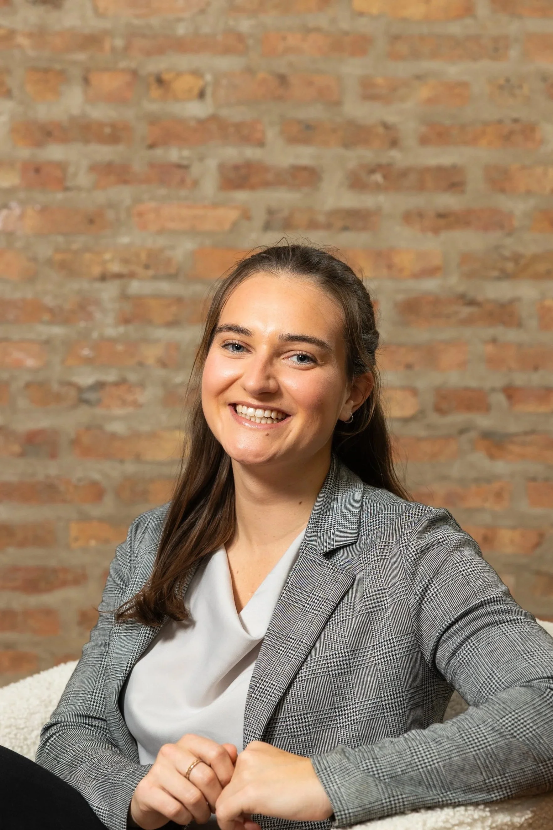 A young woman with brown hair, smiling, sitting in front of a brick wall. She is wearing a gray plaid blazer over a white blouse.