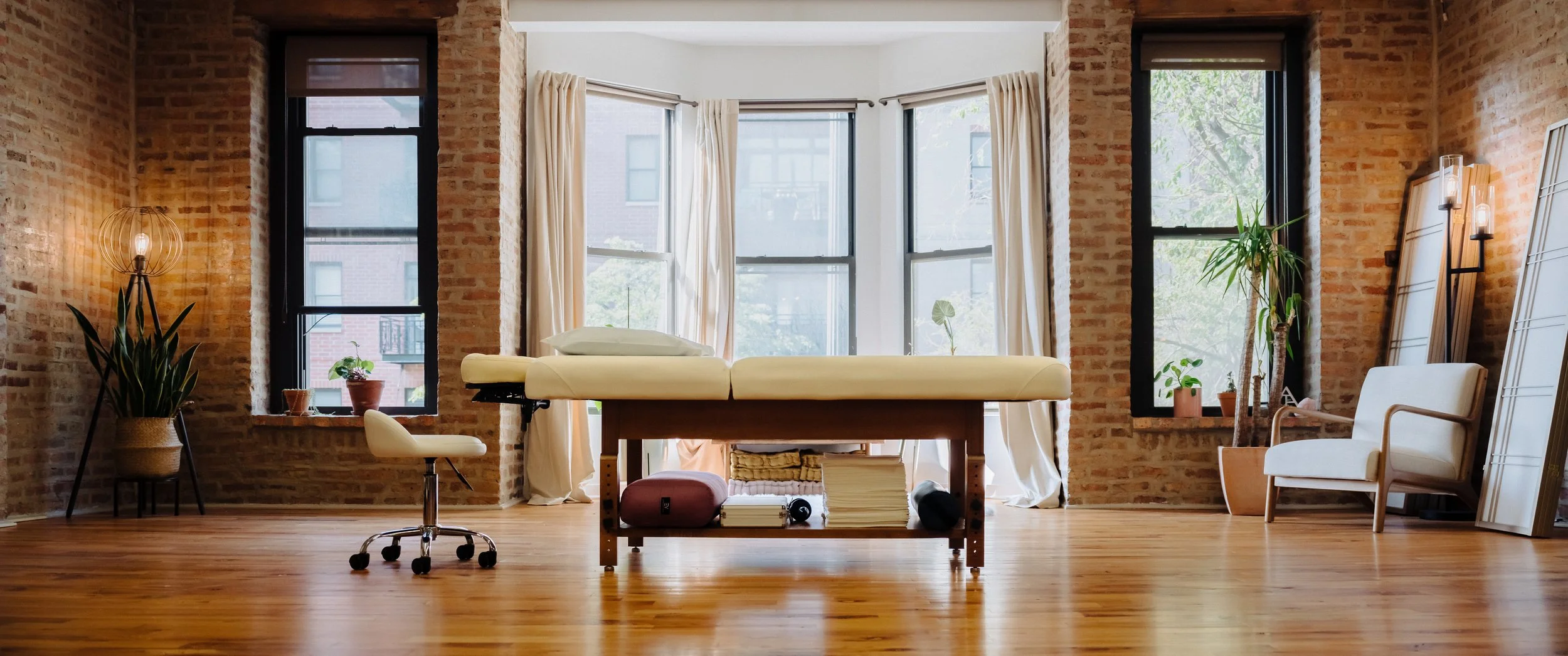 A well-lit room with large windows, brick walls, and wooden flooring. There is an exam table in the center, surrounded by plants and a chair.