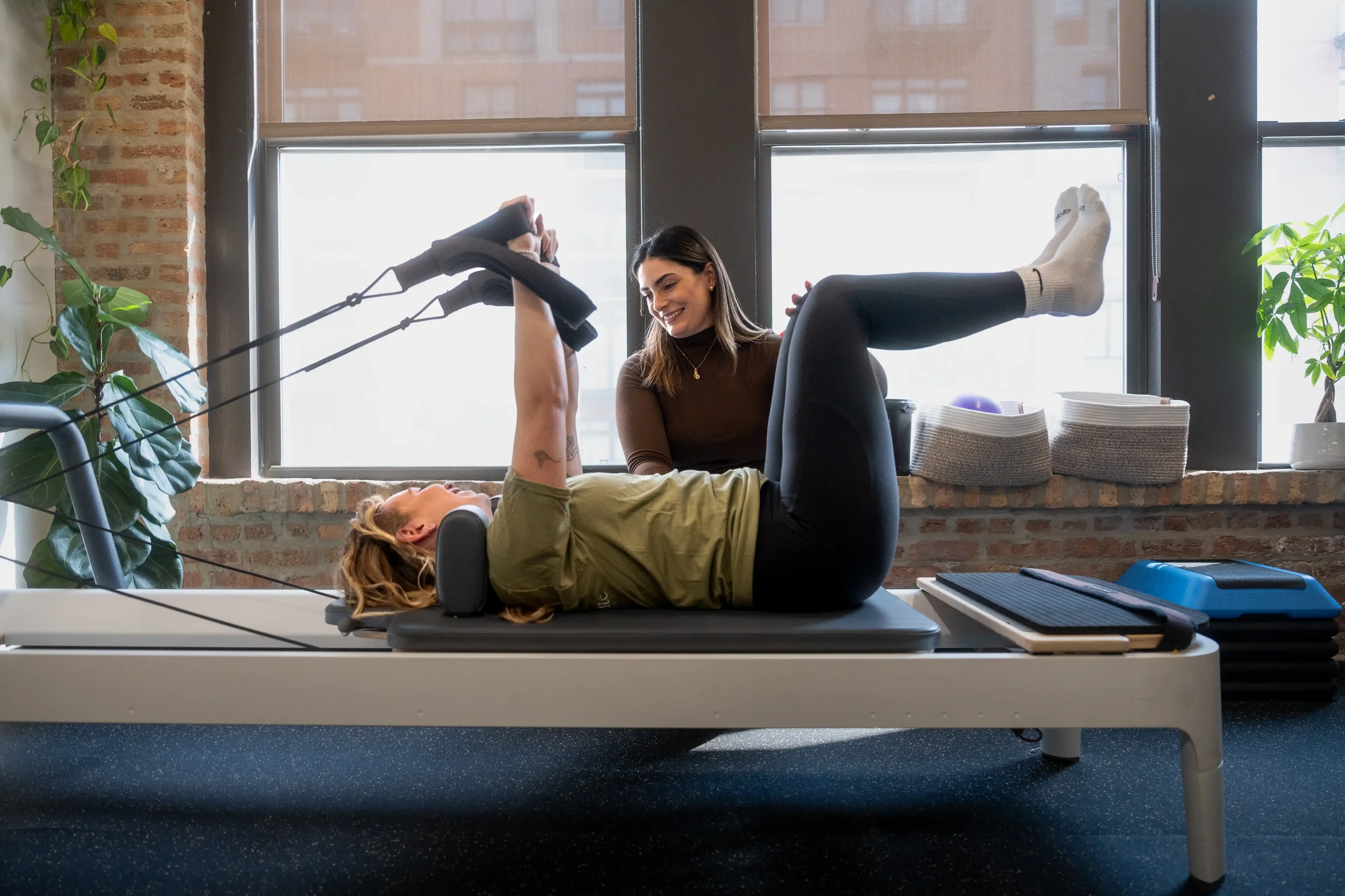 A person lying on a pilates reformer in a reverse tabletop position with her physical therapist sitting behind her. The space is bright with large windows and decorated with plants.