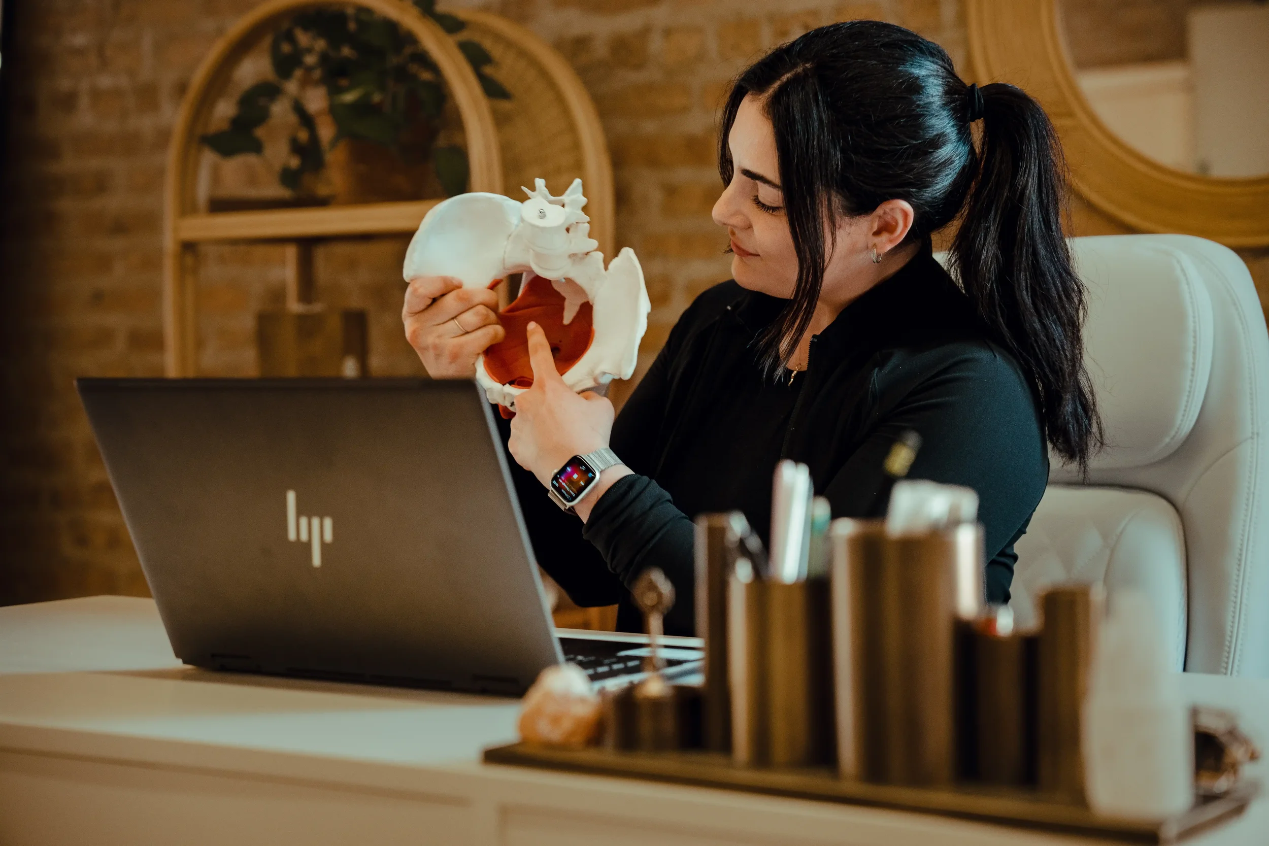 A woman holding up an anatomical model of the pelvis in front of a laptop as she works with a patient during a virtual appointment.
