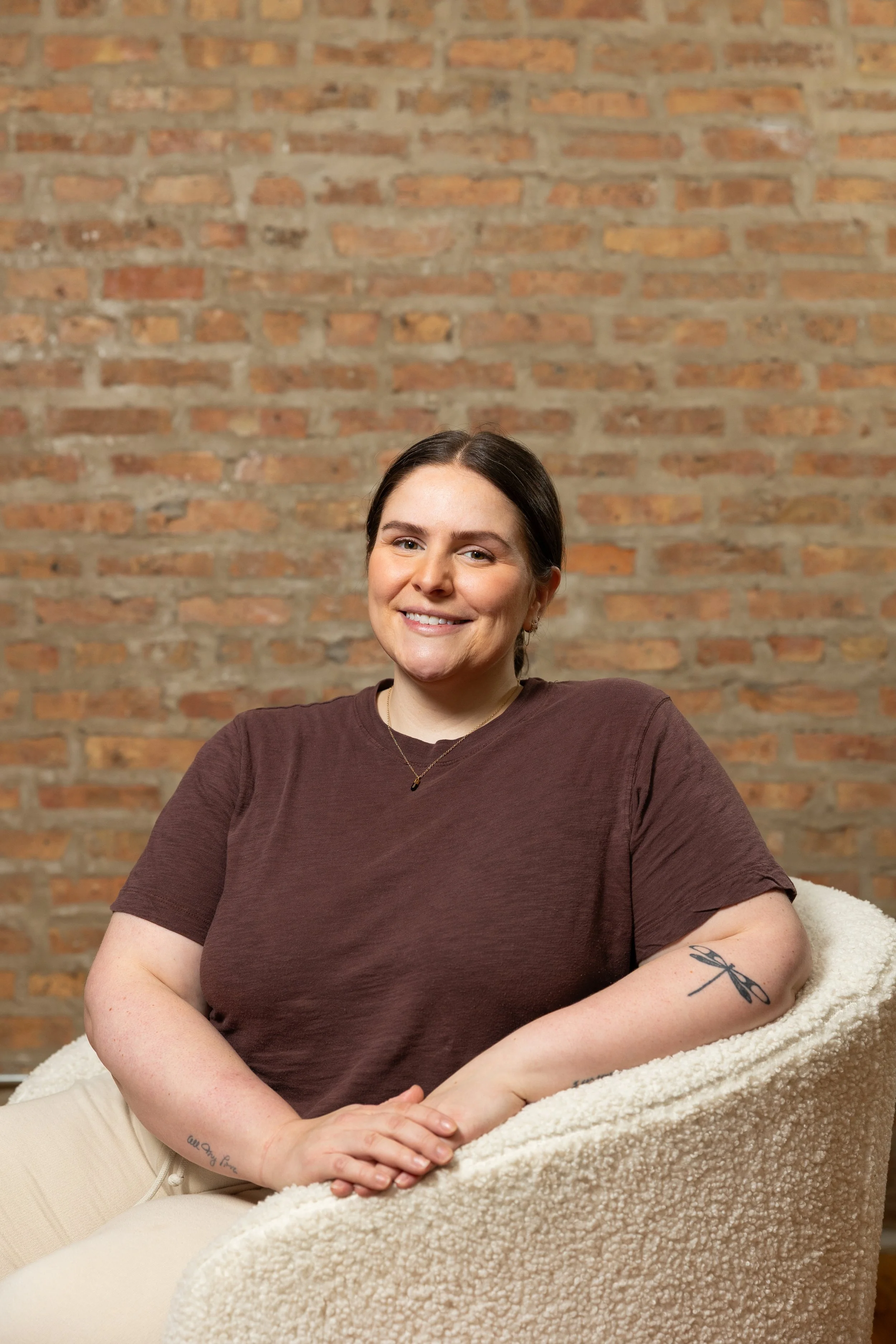 A woman with dark hair sitting on a cream-colored bean bag chair, smiling, against a brick wall background, wearing a brown t-shirt, beige pants, and a necklace, with a tattoo of a dragonfly on her arm.