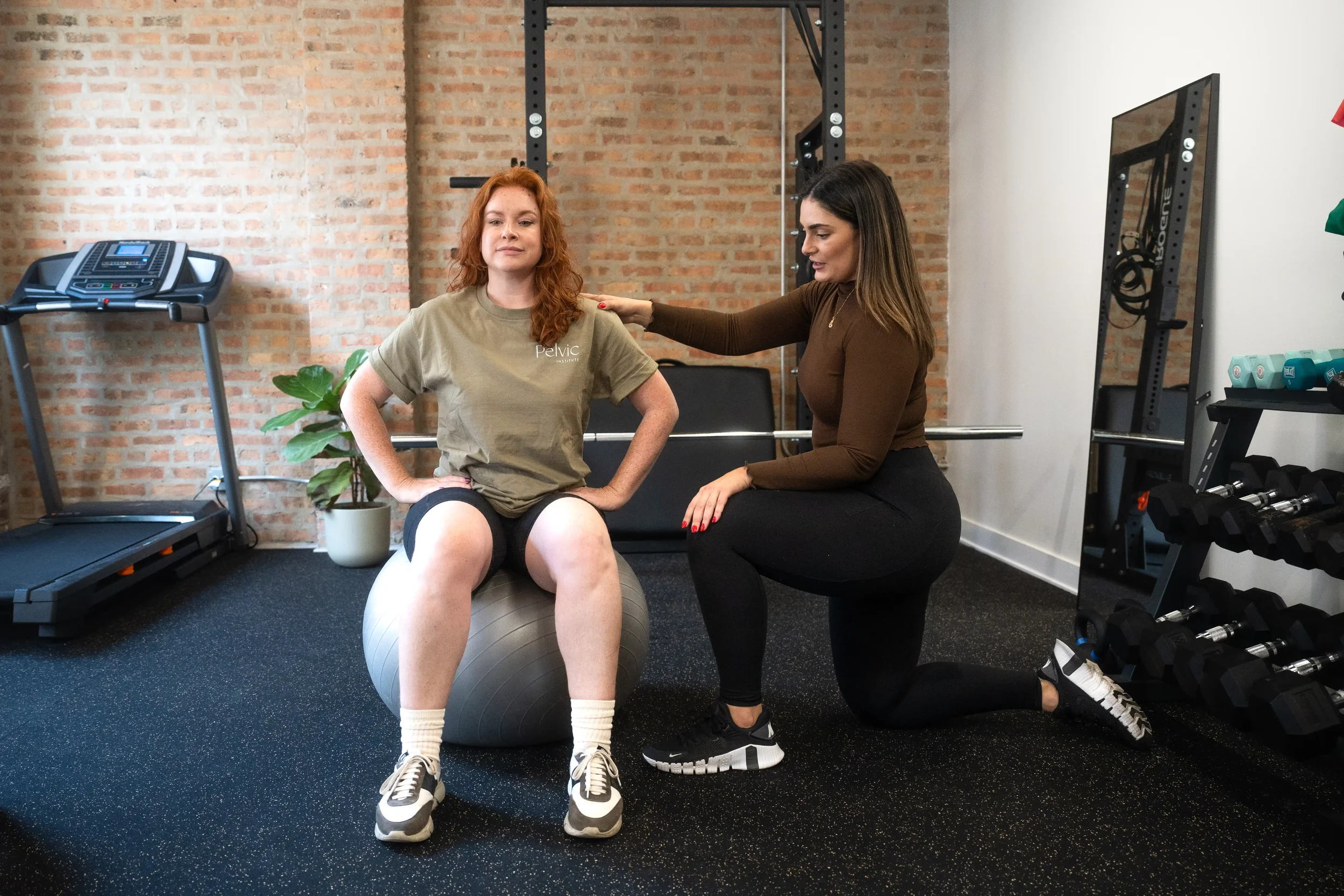 A woman with red hair sitting on an exercise ball during a fitness session with a trainer in a physical therapy office, brick wall background, treadmills, mirror, and workout equipment visible.
