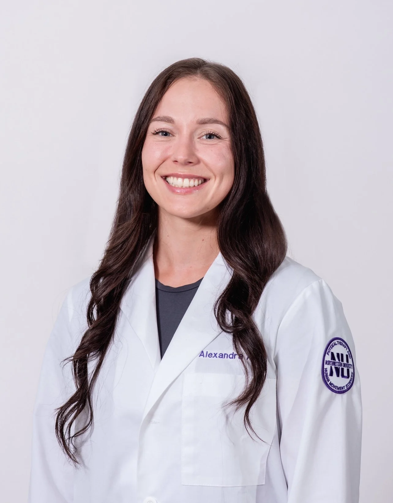 A young woman with long, wavy dark hair and light skin, smiling, wearing a white medical coat with a name tag reading 'Alexandra'.