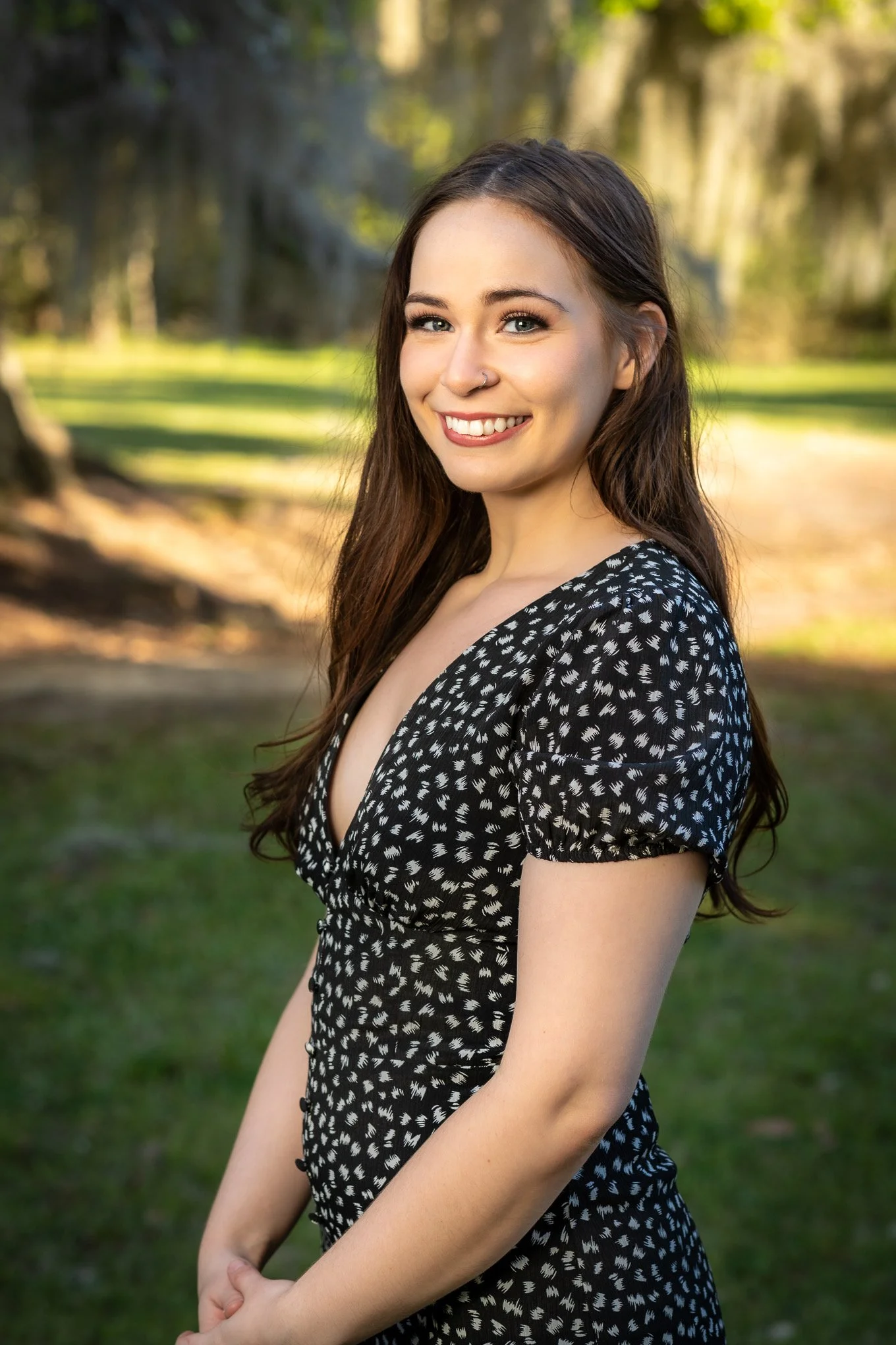 A young woman with long brown hair, a nose piercing, and bright blue eyes standing outdoors in a park-like setting, smiling at the camera, wearing a black and white polka dot dress.