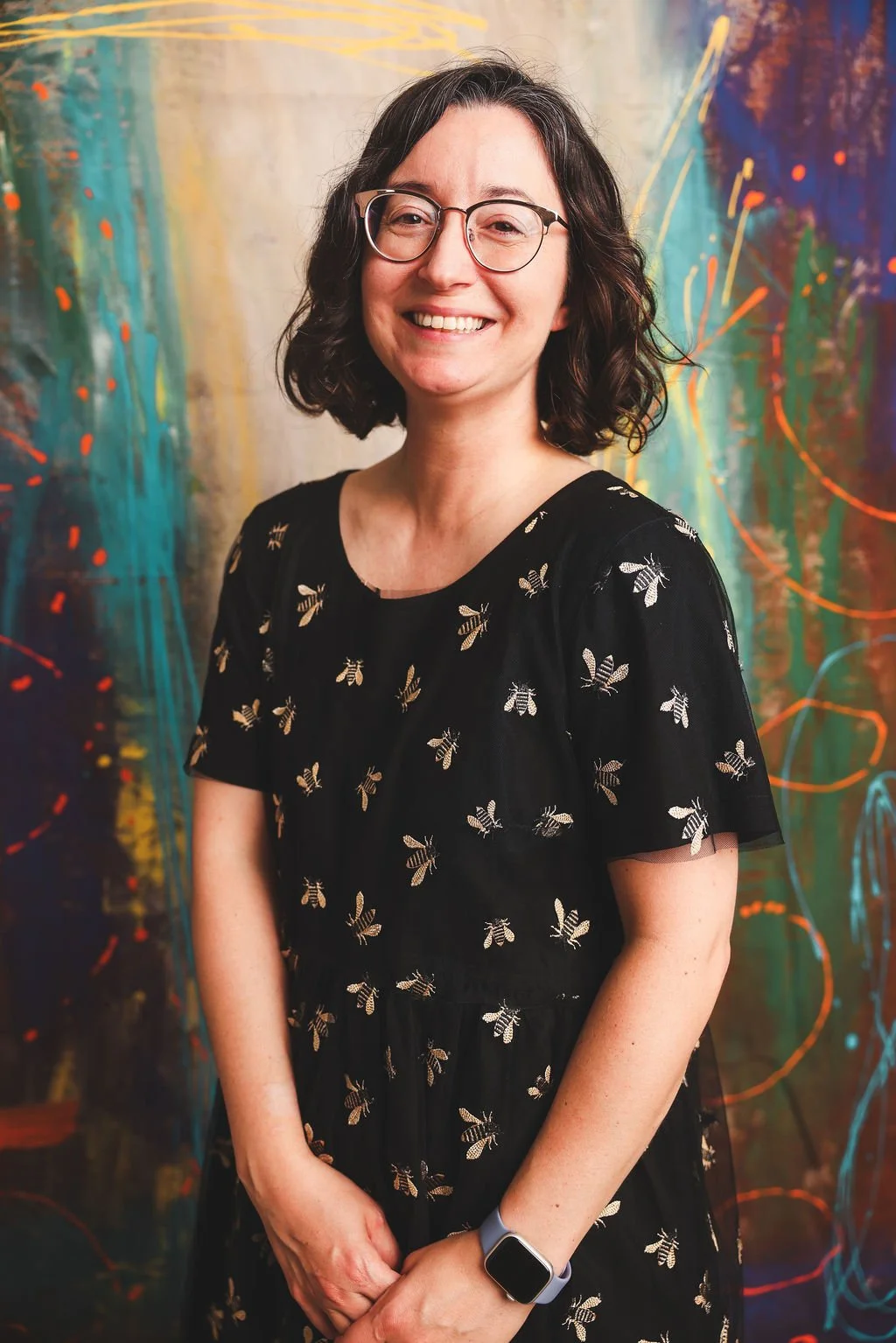 Rebecca Huval, grief therapist, smiles in front of a vibrant abstract painting with a green background and orange paint splatters. Rebecca works with disenfranchised grief, including chronic illness, infertility, and pet loss.