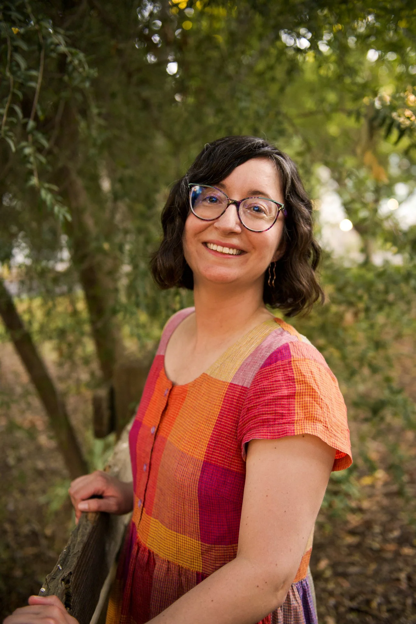 Rebecca Huval, grief therapist, leans toward the viewer and smiles. She's holding onto a fence in the Sacramento State Arboretum.
