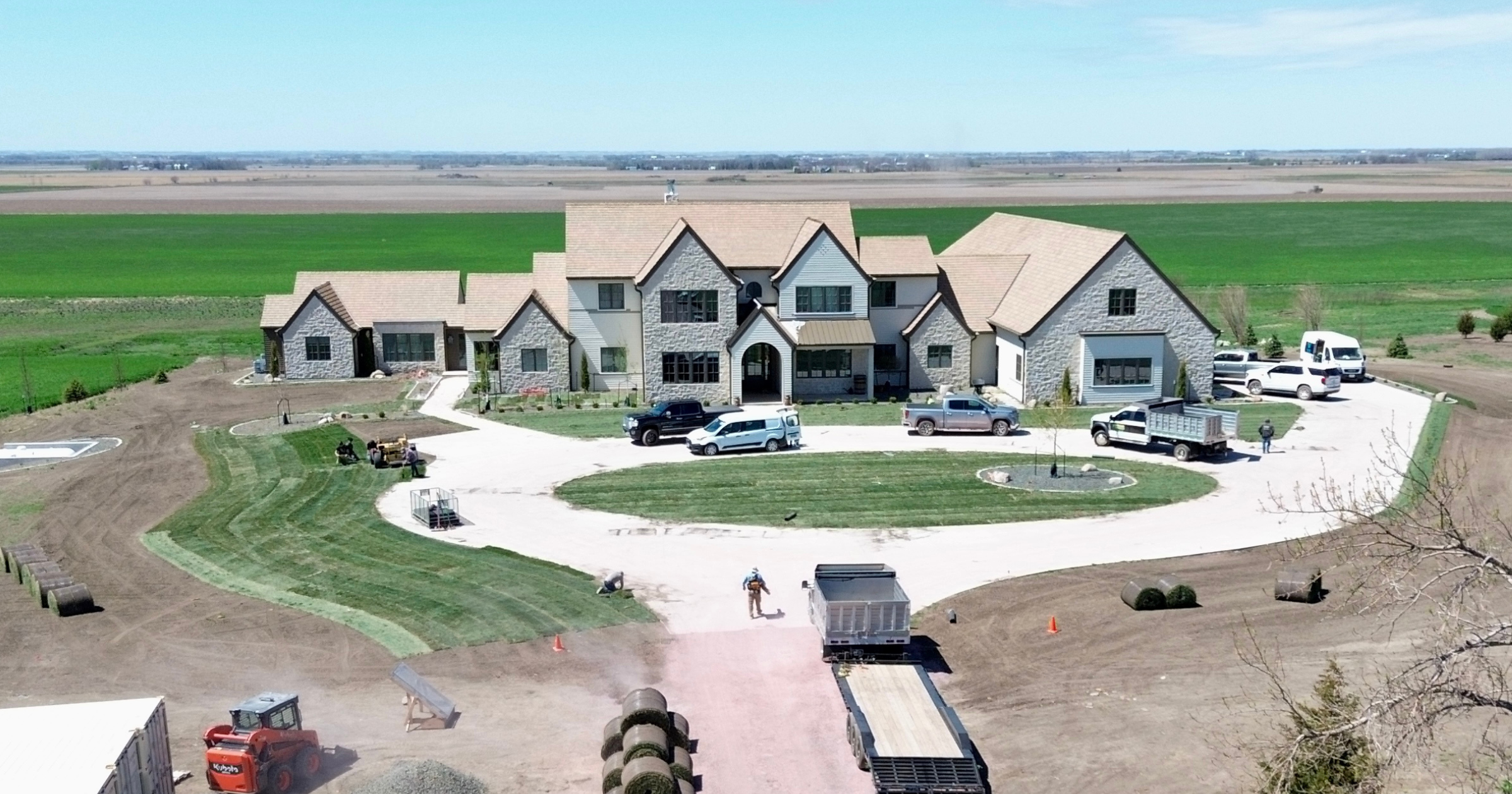 A large house under construction in a rural area with multiple vehicles parked in the driveway and construction equipment around the yard.