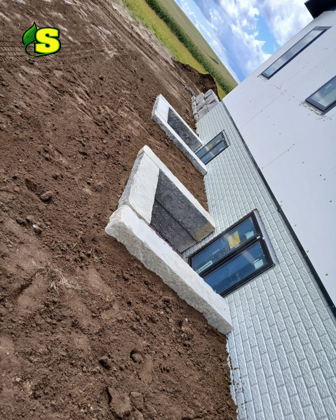 Construction site with stone and brick landscape walls being built next to a white brick building with large windows, soil in the foreground, and a cloudy sky.