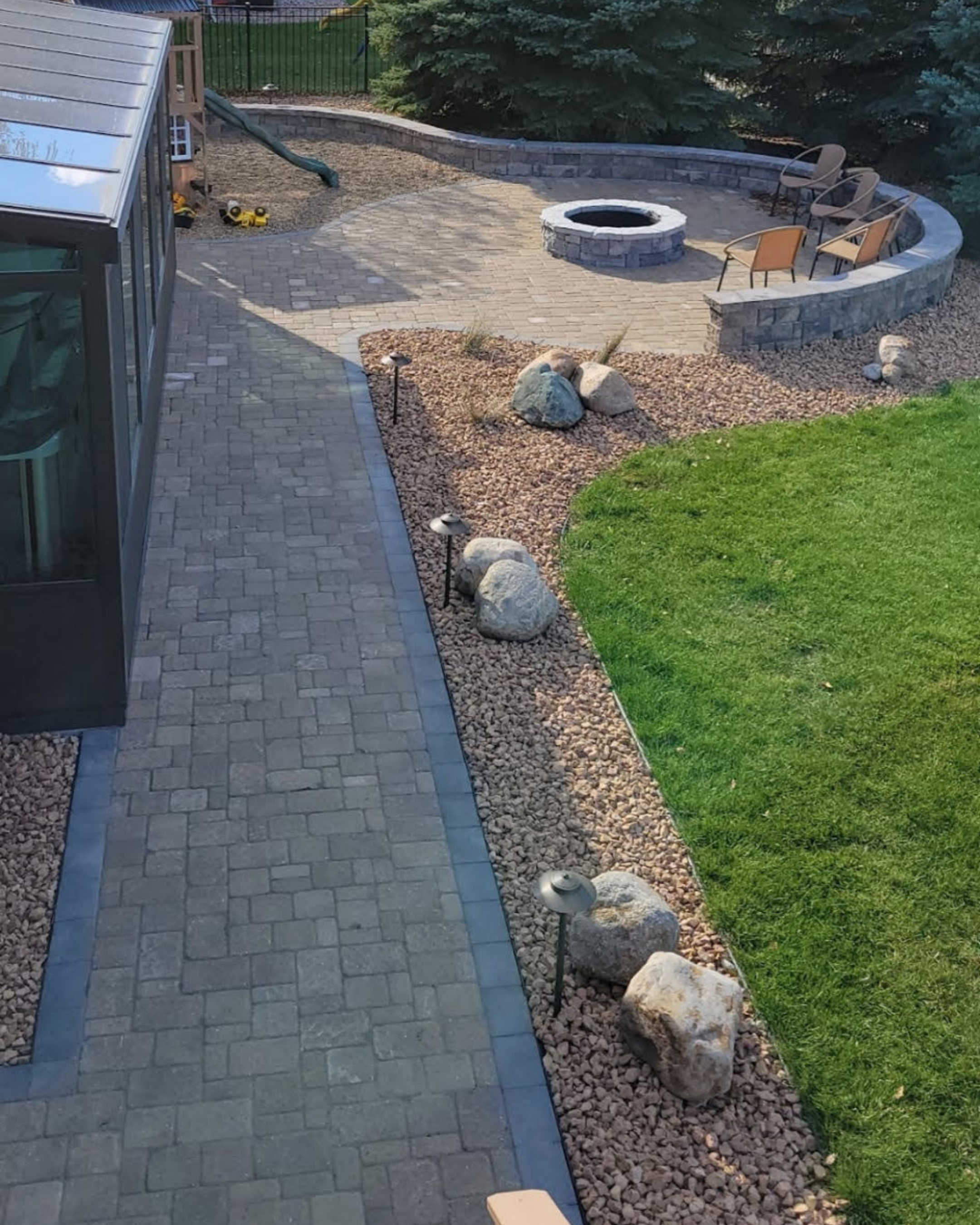 Backyard with a paved patio, a fire pit surrounded by four chairs, large rocks, a rock garden, and a slide, with a greenhouse on the left.