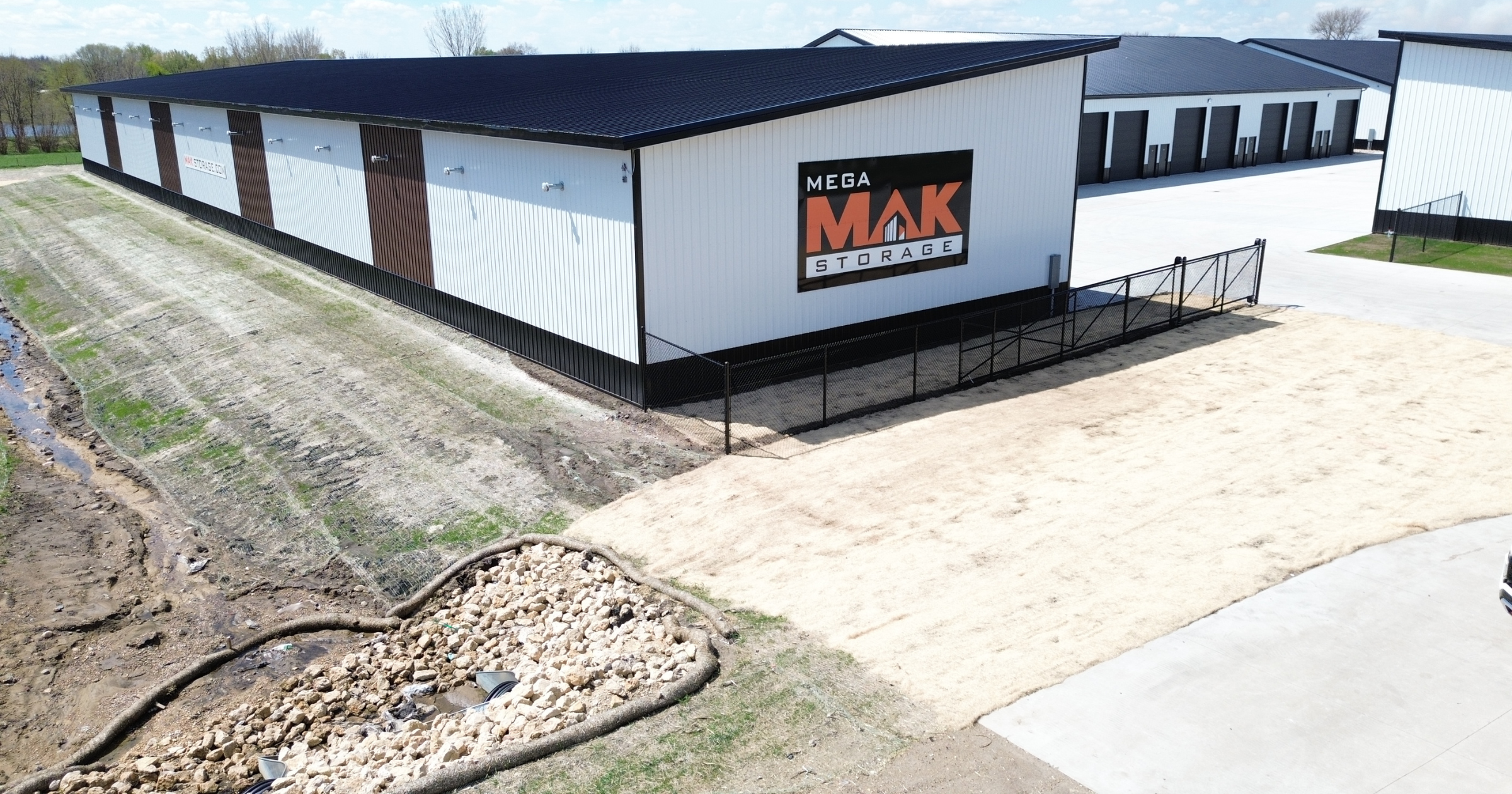 A large storage building with a sign that reads 'Mega MAK Storage'. The building has black and white exterior walls, a black roof, and multiple roll-up doors. The area in front of the building includes a paved driveway, fencing, and a sandy patch of ground. There are some trees and a body of water in the background.
