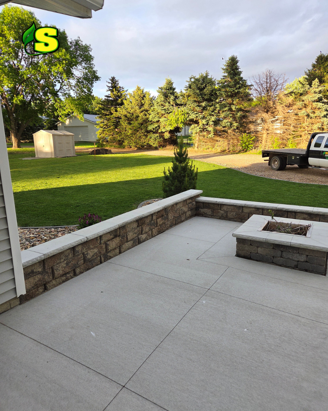 View from a porch with a concrete floor and a brick and stone railing, overlooking a green lawn with trees, a garden shed, and a pickup truck parked on gravel driveway in the background