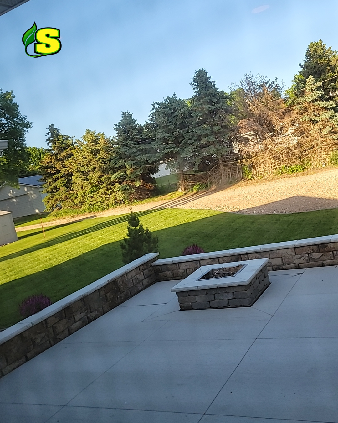 View of backyard patio with stone firepit, green lawn, trees, and blue sky.