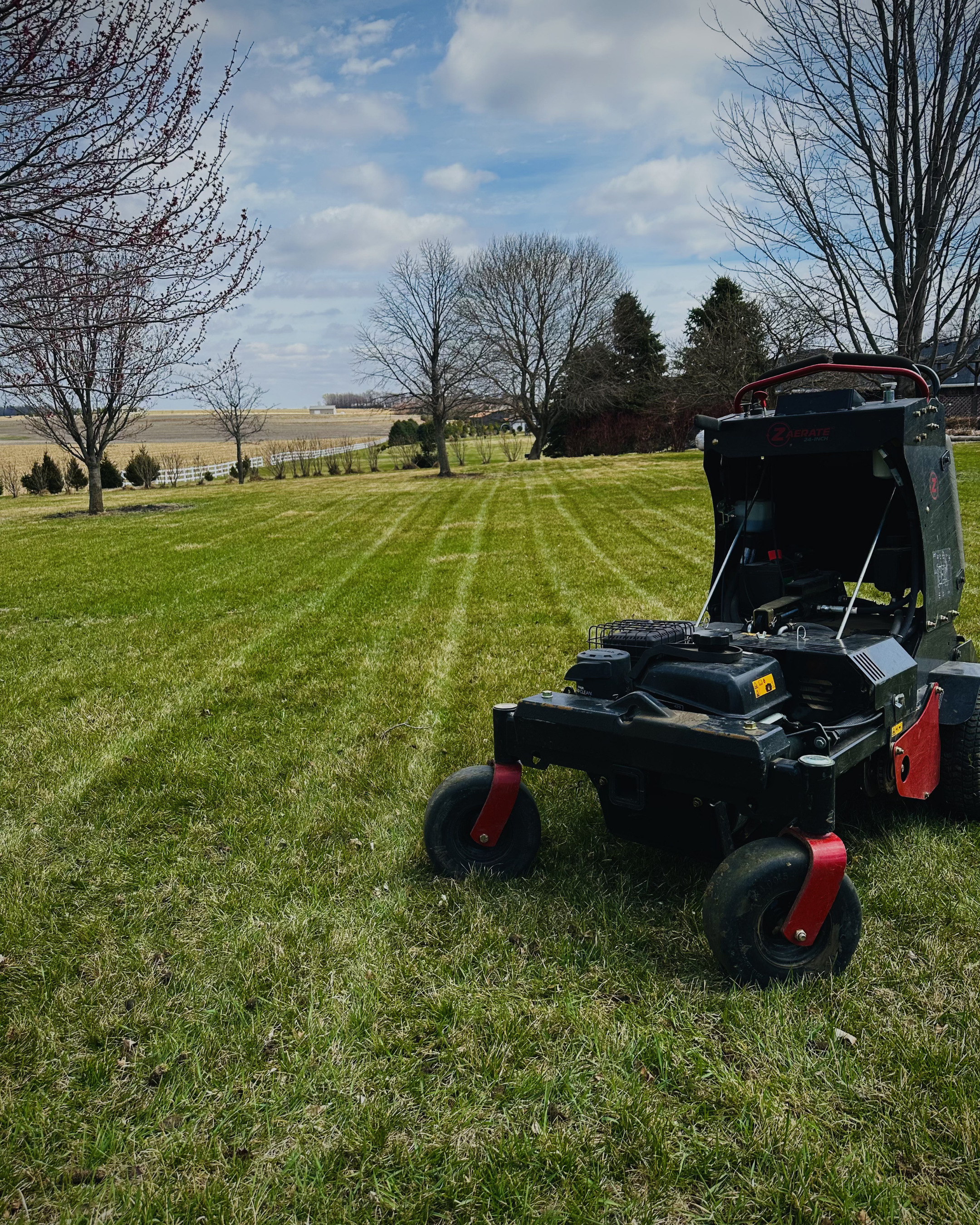 A lawn with freshly cut grass, several leafless trees, and a corner of a piece of outdoor equipment on the right side, with a partly cloudy sky overhead and a distant farmhouse or barn in the background.