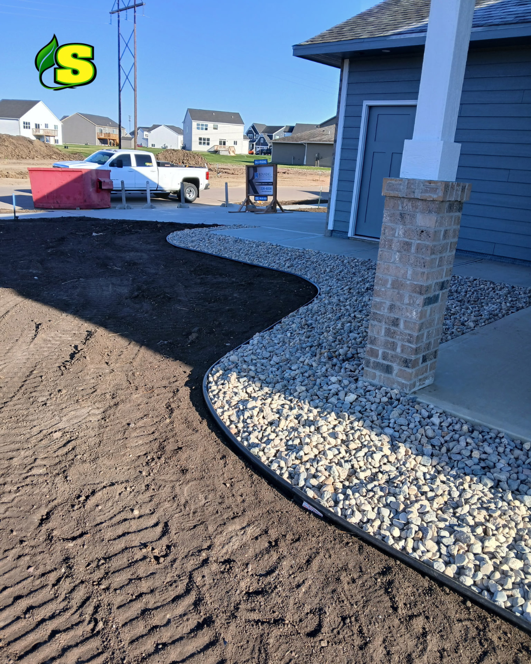 View of a house exterior with a brick pillar, gravel landscaping, and a sidewalk. In the background, there is a white pickup truck and a large red construction container, suggesting ongoing construction or landscaping work.