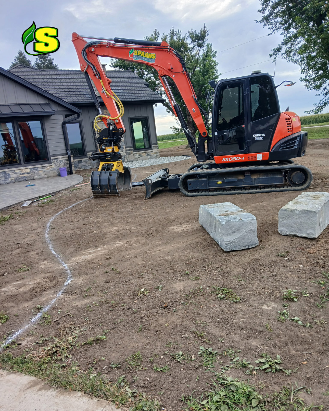 A small construction excavator working on a dirt yard in front of a house, with two large stone blocks nearby and a curved chalk line on the ground.