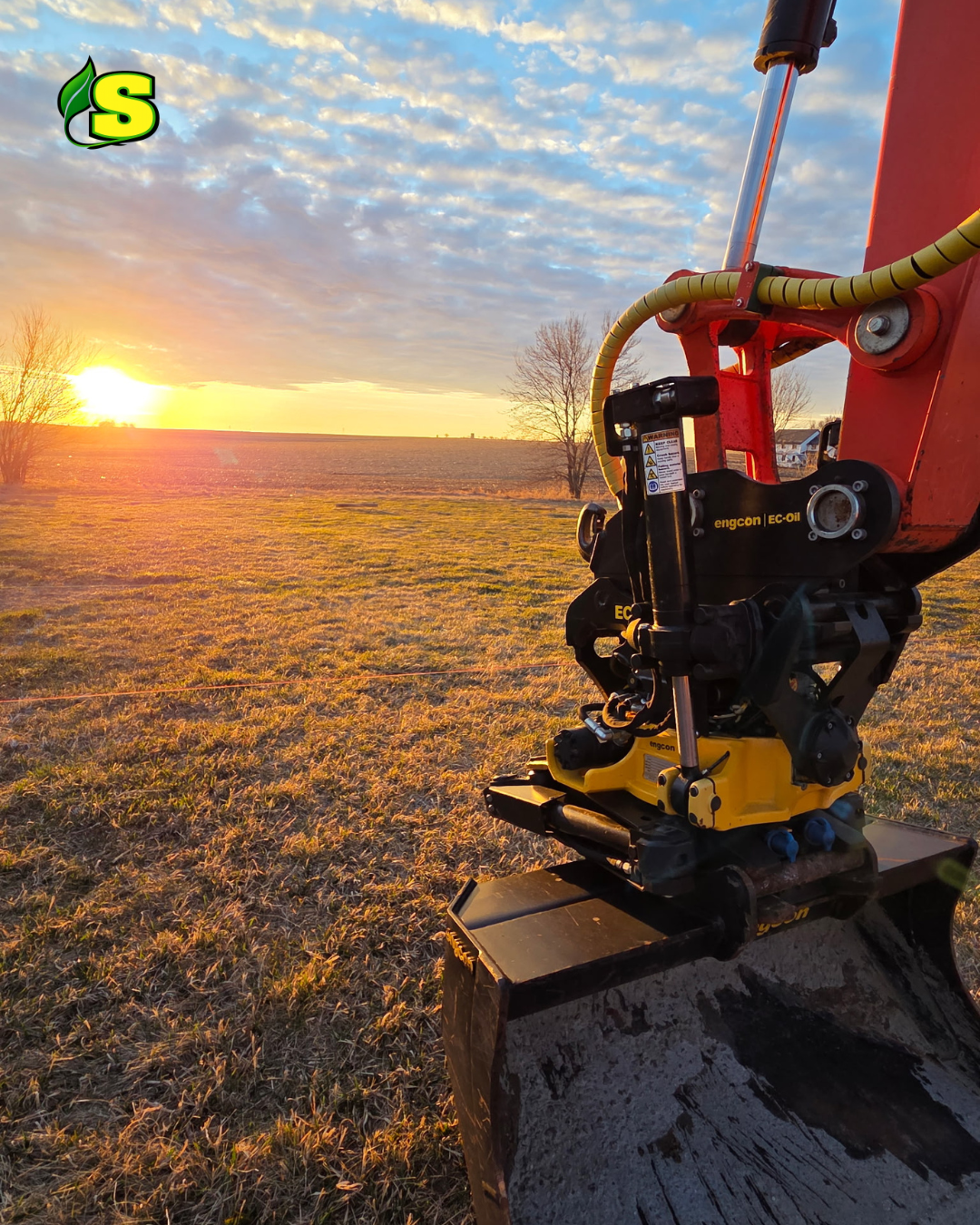 A construction or gardening machine's hydraulic arm with yellow tubing and black components in a field at sunset, with a partly cloudy sky.