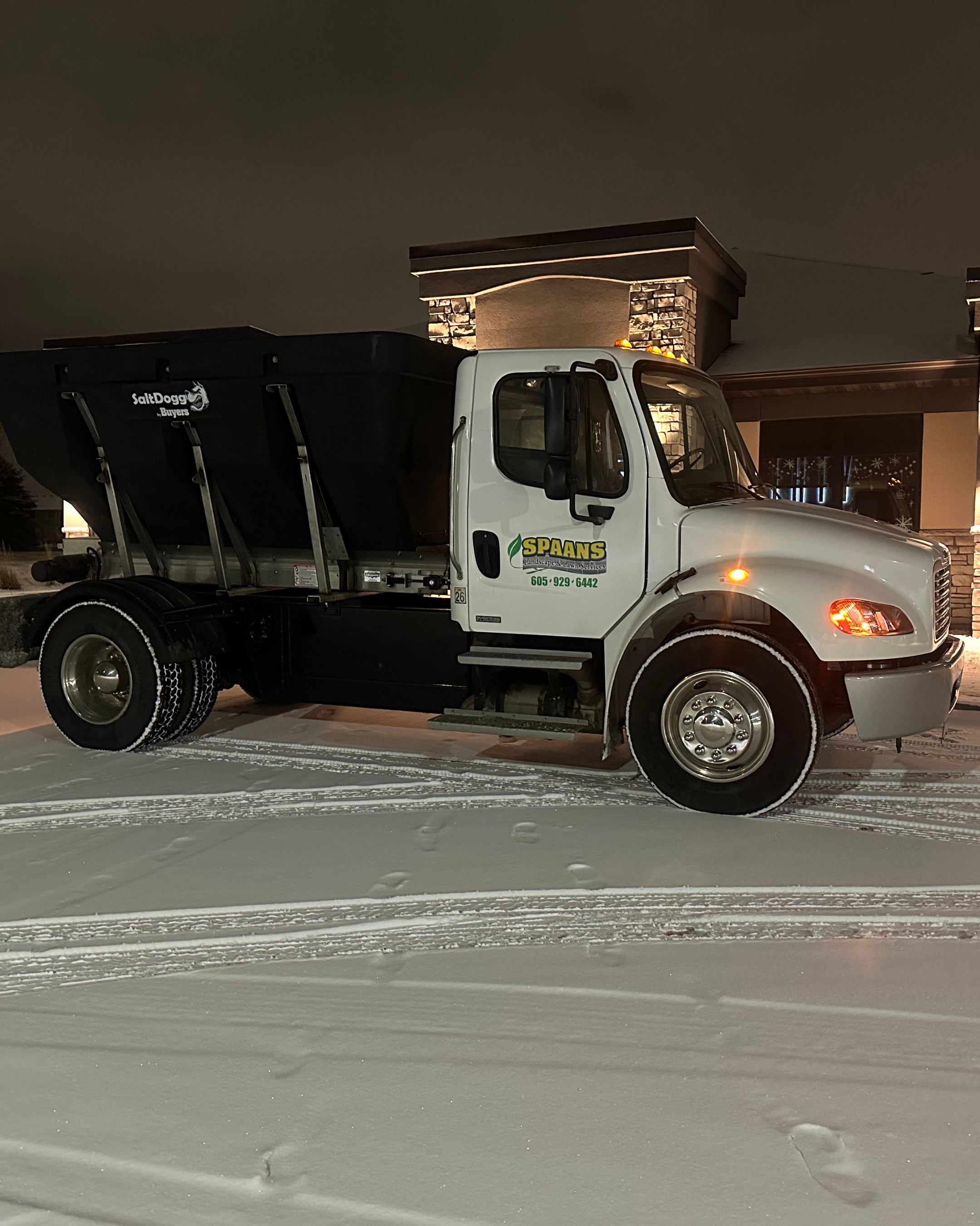 A white truck with a black cargo area, parked on a snow-covered surface at night, belongs to Spaans Landscape & Lawn Services, with contact number 605-929-6442, in front of a building with stone accents and lit windows.
