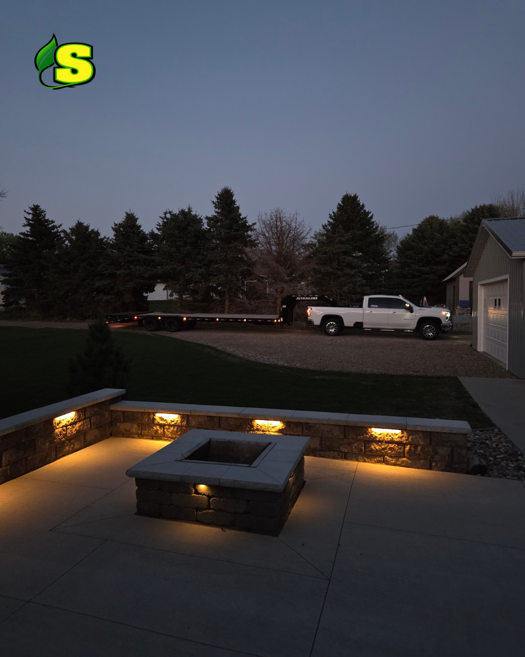 Nighttime view of a backyard patio with built-in firepit and stone wall, illuminated by warm outdoor lights, a white truck towing a trailer parked on the driveway, trees and a shed in the background under a darkening sky.