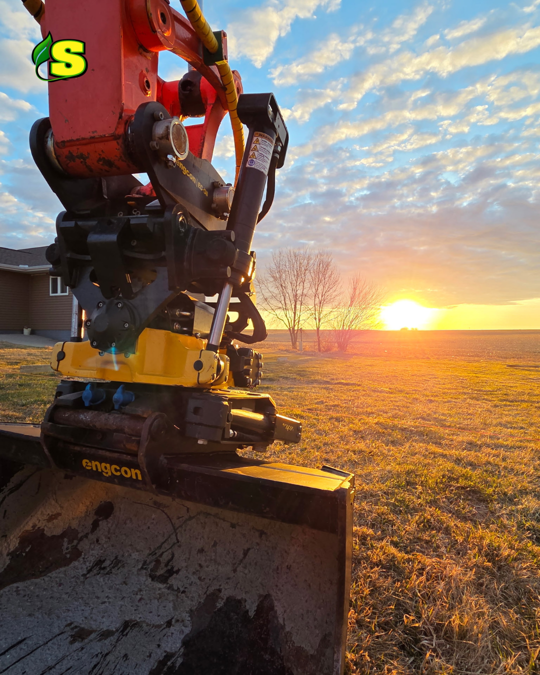 Close-up of an excavator attachment with a sunset and partly cloudy sky in the background.
