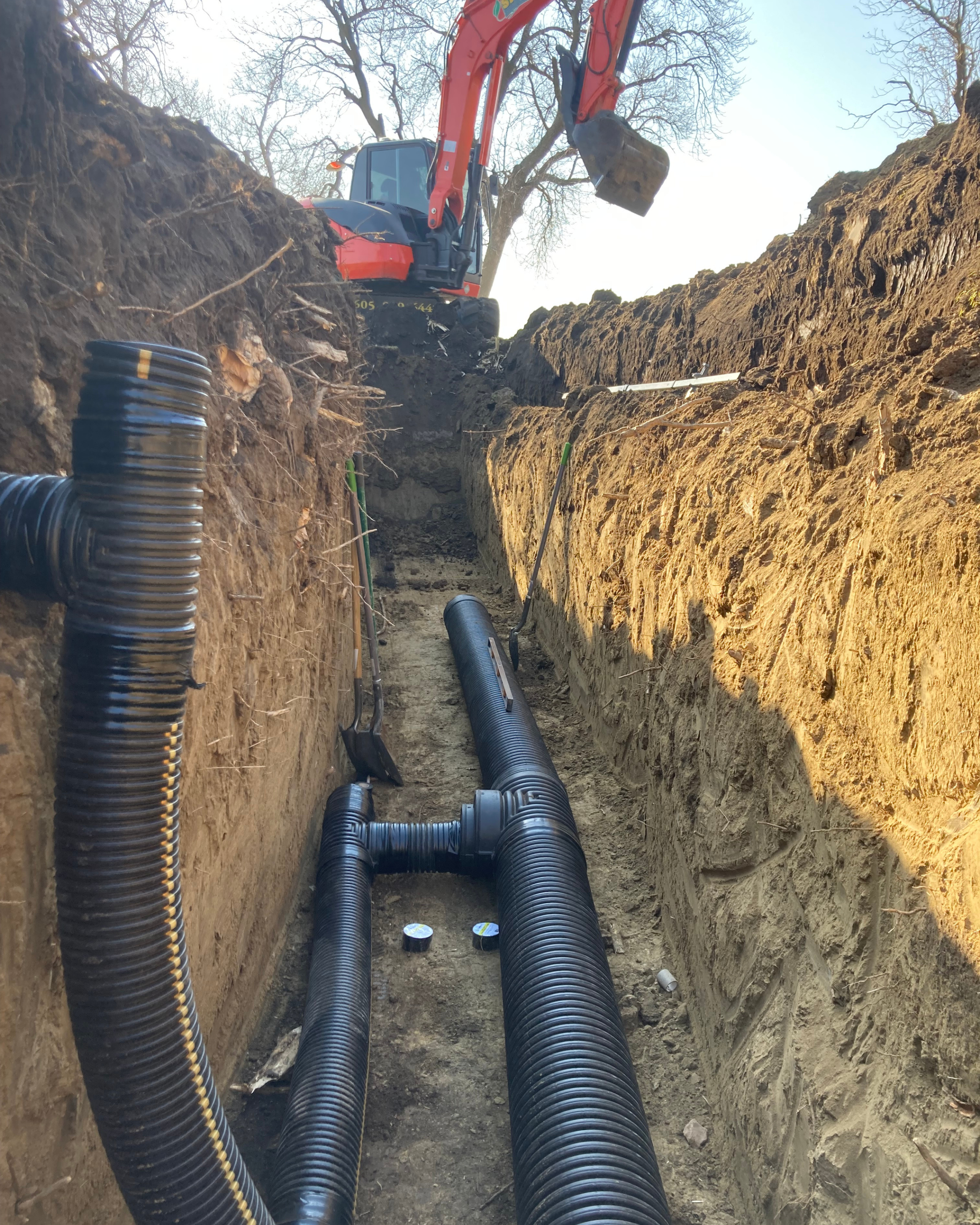 View of a trench with black corrugated pipes installed for underground utilities, with a small excavator in the background.