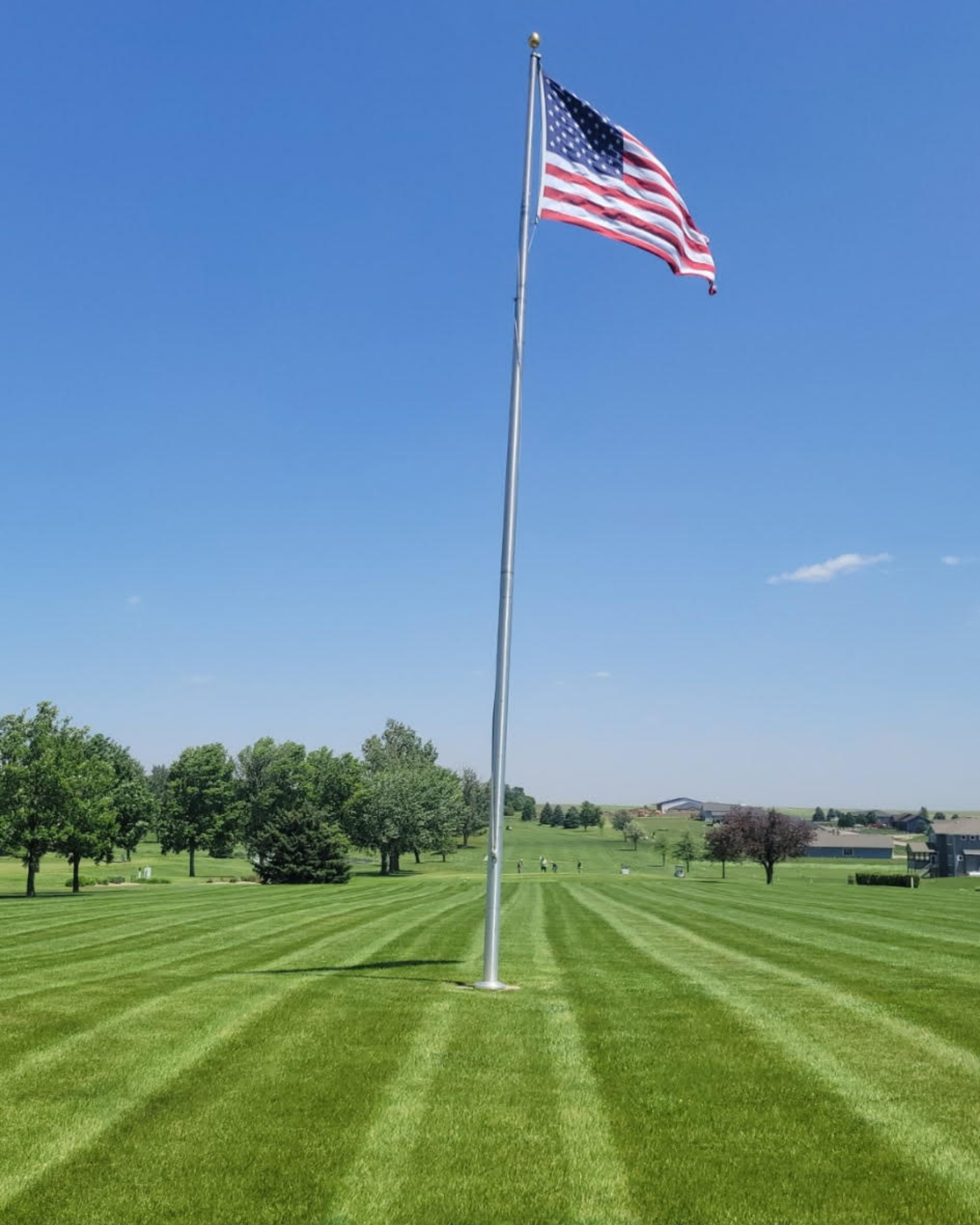 American flag flying on a flagpole in a well-maintained park with green lawn, trees, and distant buildings under a clear blue sky.