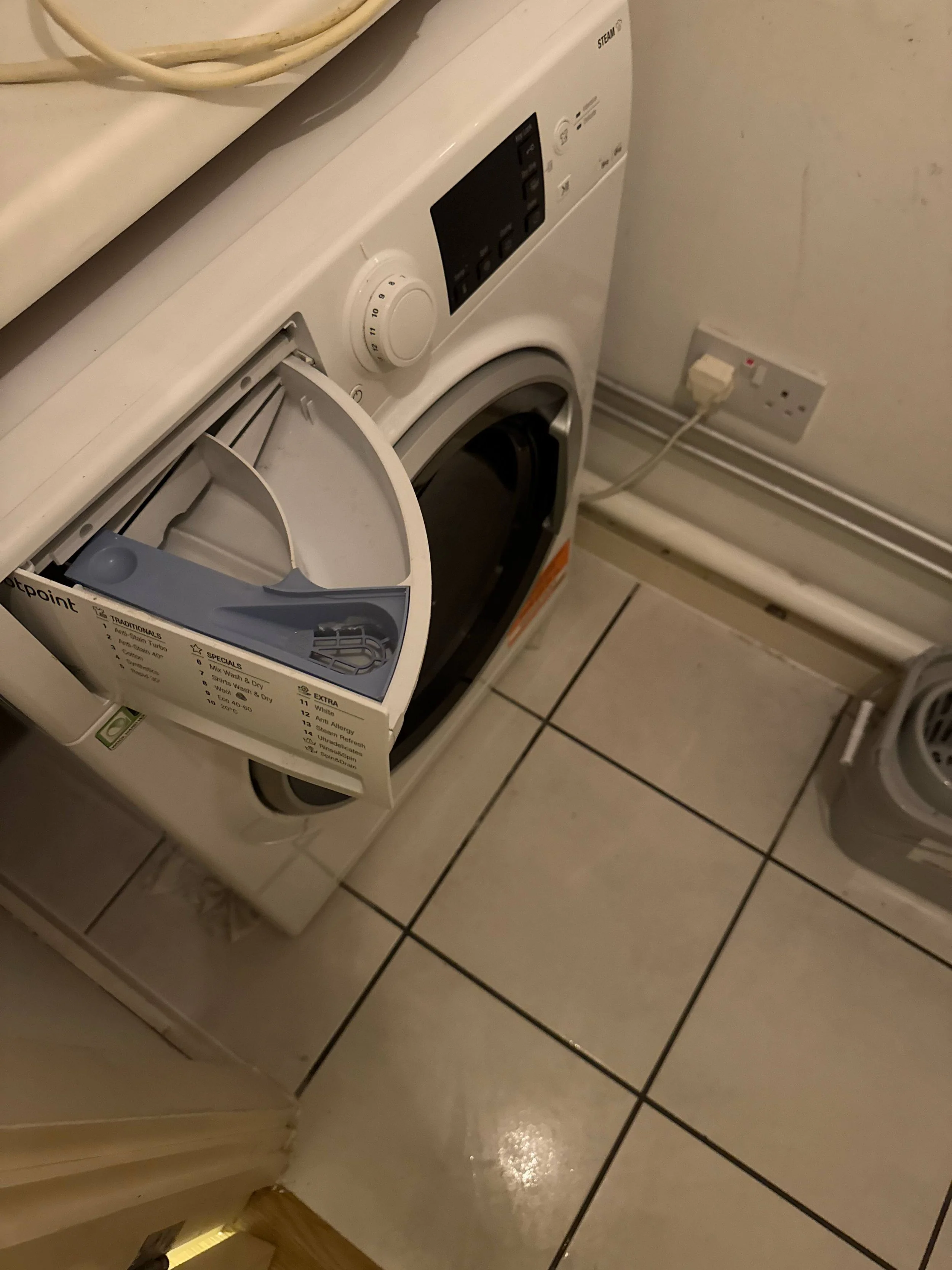 A front-loading washing machine with the door open, placed on a tiled floor next to a wall with an electrical outlet.