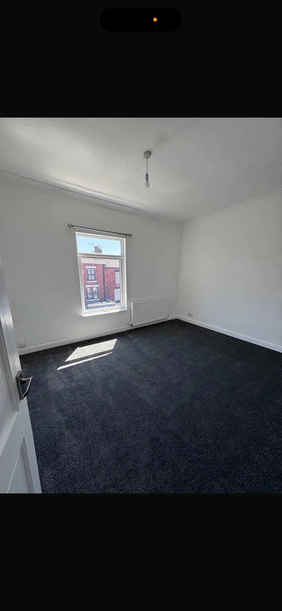 Empty white room with a window, dark carpeted floor, and a radiator, sunlight coming through the window.