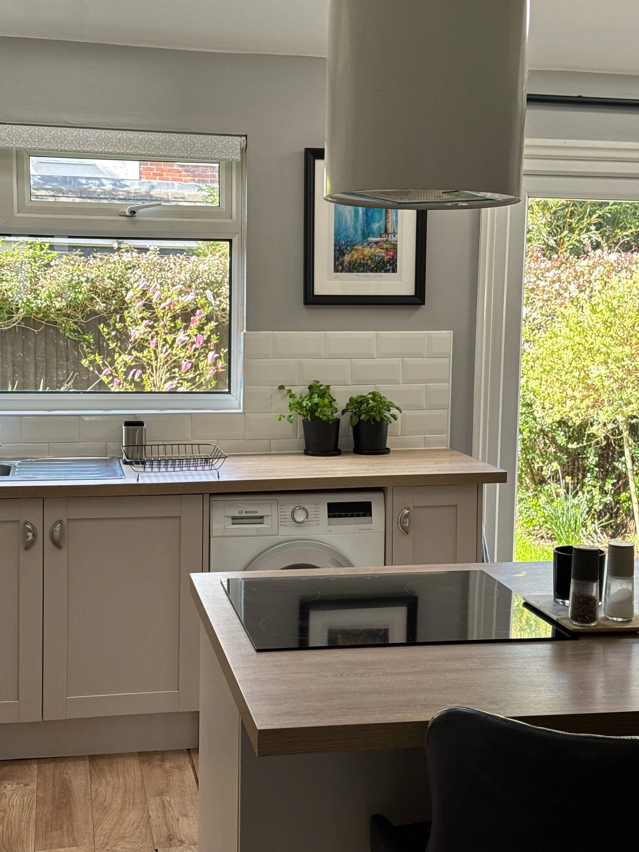 A modern kitchen with a light-colored wooden countertop, white cabinetry, a black induction cooktop, and a washing machine underneath the counter. Two small potted plants are on the countertop, and a framed painting hangs on the wall. There's a window letting in natural light and a sliding door with a view of a garden outside.