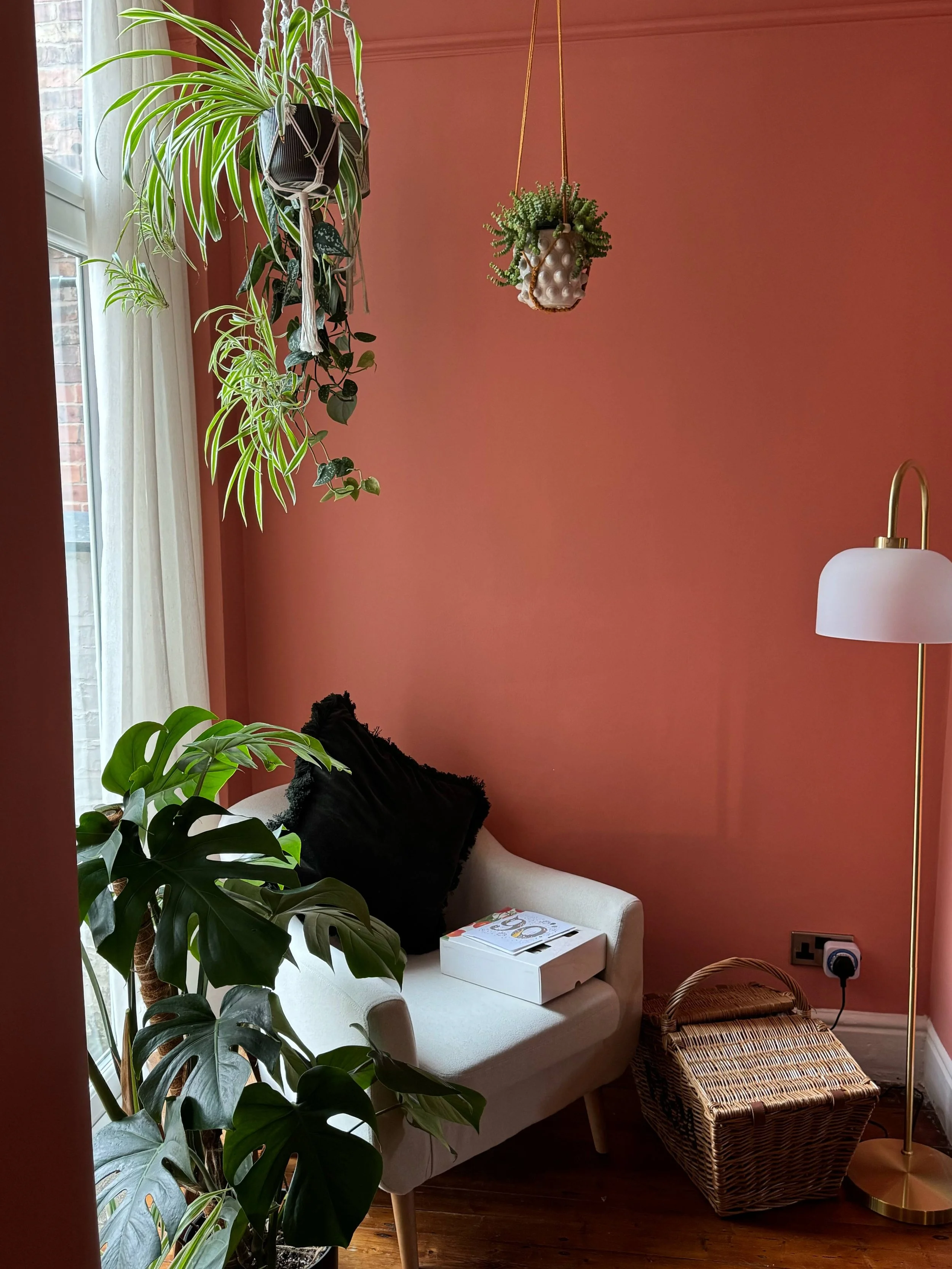 A cozy corner with a beige armchair, black pillow, and a window with cream curtains. Hanging plants adorn the corner, and a wicker basket and white box sit on the floor. A gold floor lamp is nearby, and the wall is painted a warm terracotta color.