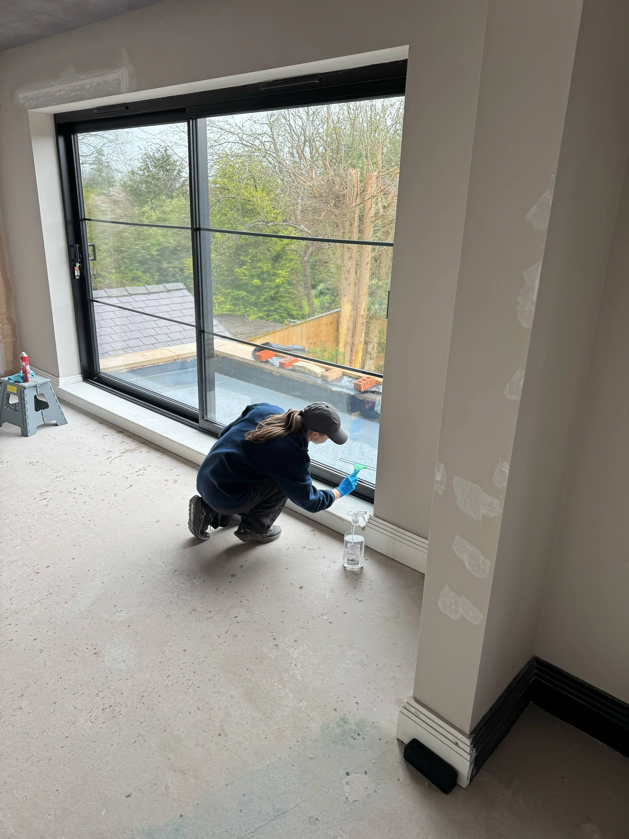 Person kneeling on floor placing window frame or window sill, with tools and spray bottle nearby, in a room under construction.