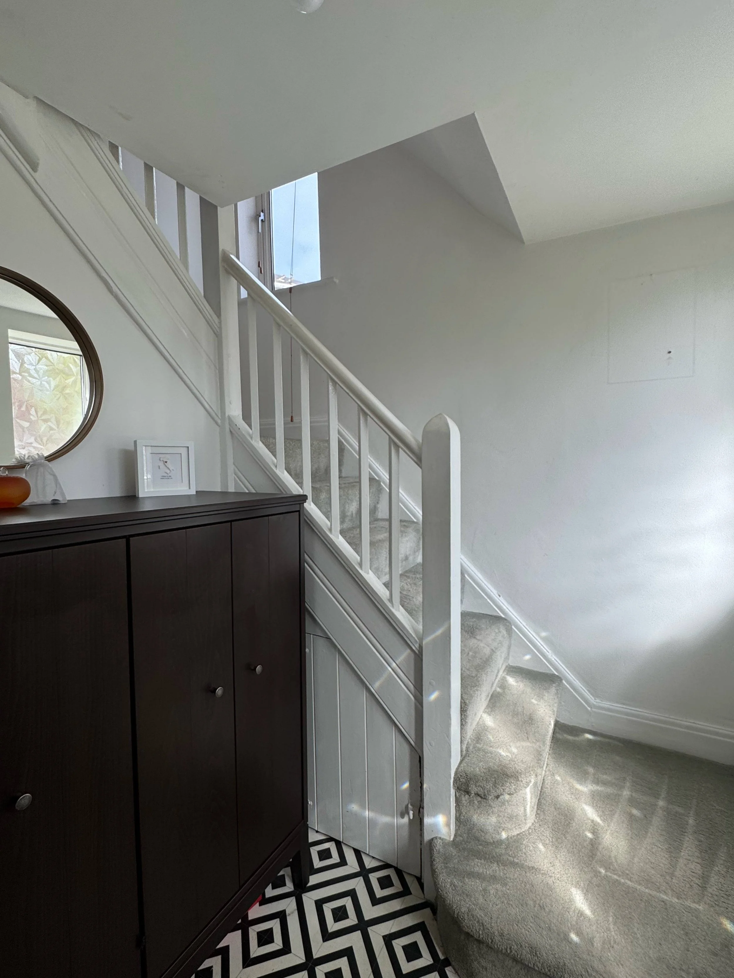Interior of a house with a staircase, a black cabinet, a round mirror, a framed picture, a table lamp, and a patterned rug, with sunlight streaming through windows.
