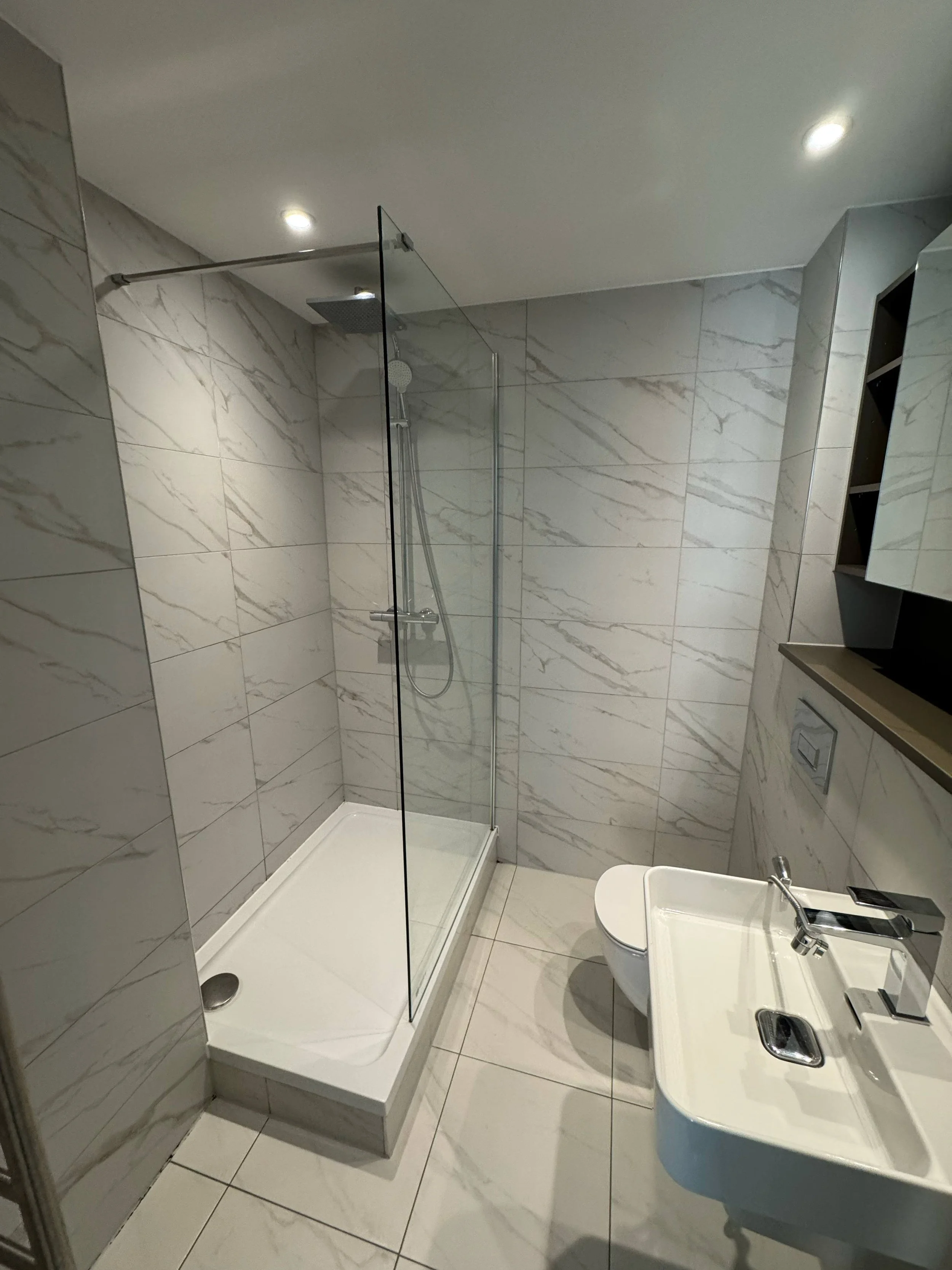 A modern bathroom featuring a glass shower stall with a rainfall showerhead, white tiled walls with gray marble veining, a white toilet, and a small white sink with a chrome faucet.
