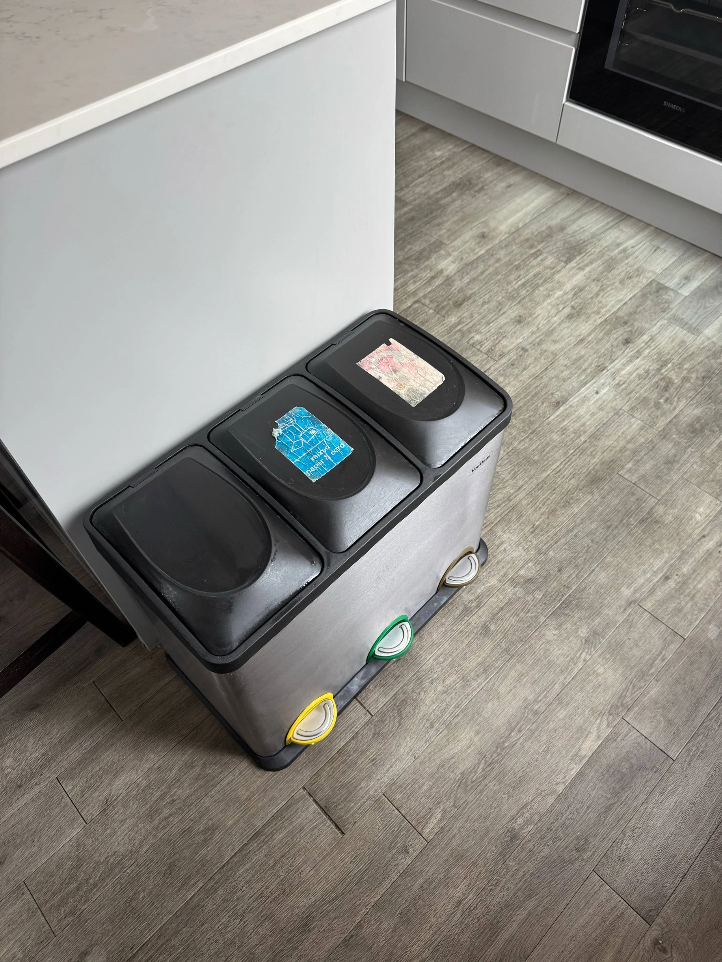 Two trash bins placed next to a white kitchen counter with a stove in the background, and a wood-patterned floor.