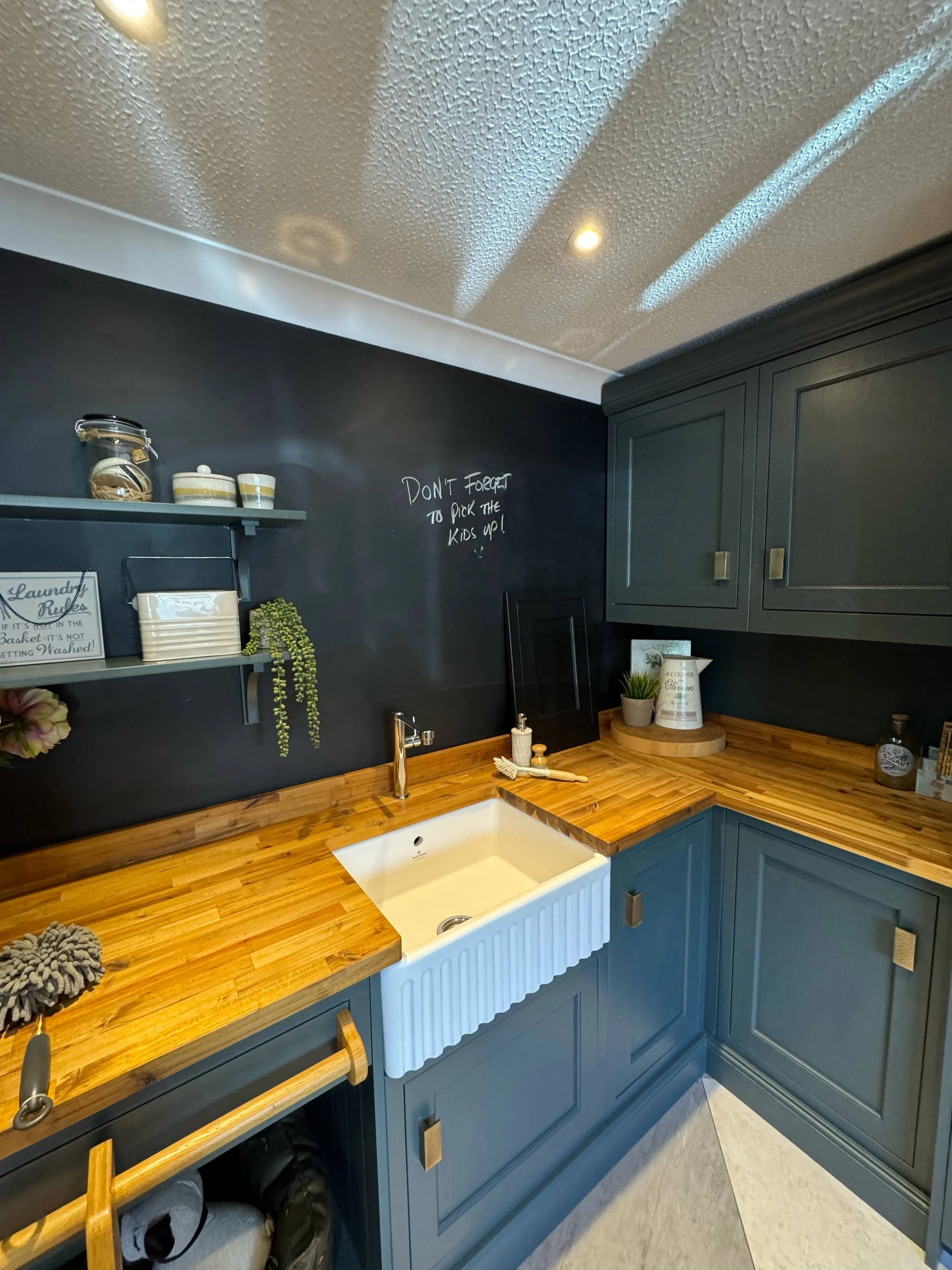 A kitchen with blue cabinets, a white farmhouse sink, wooden countertops, and dark walls. There are shelves with decorative containers and a chalkboard with a note. The ceiling has a textured finish and recessed lighting.
