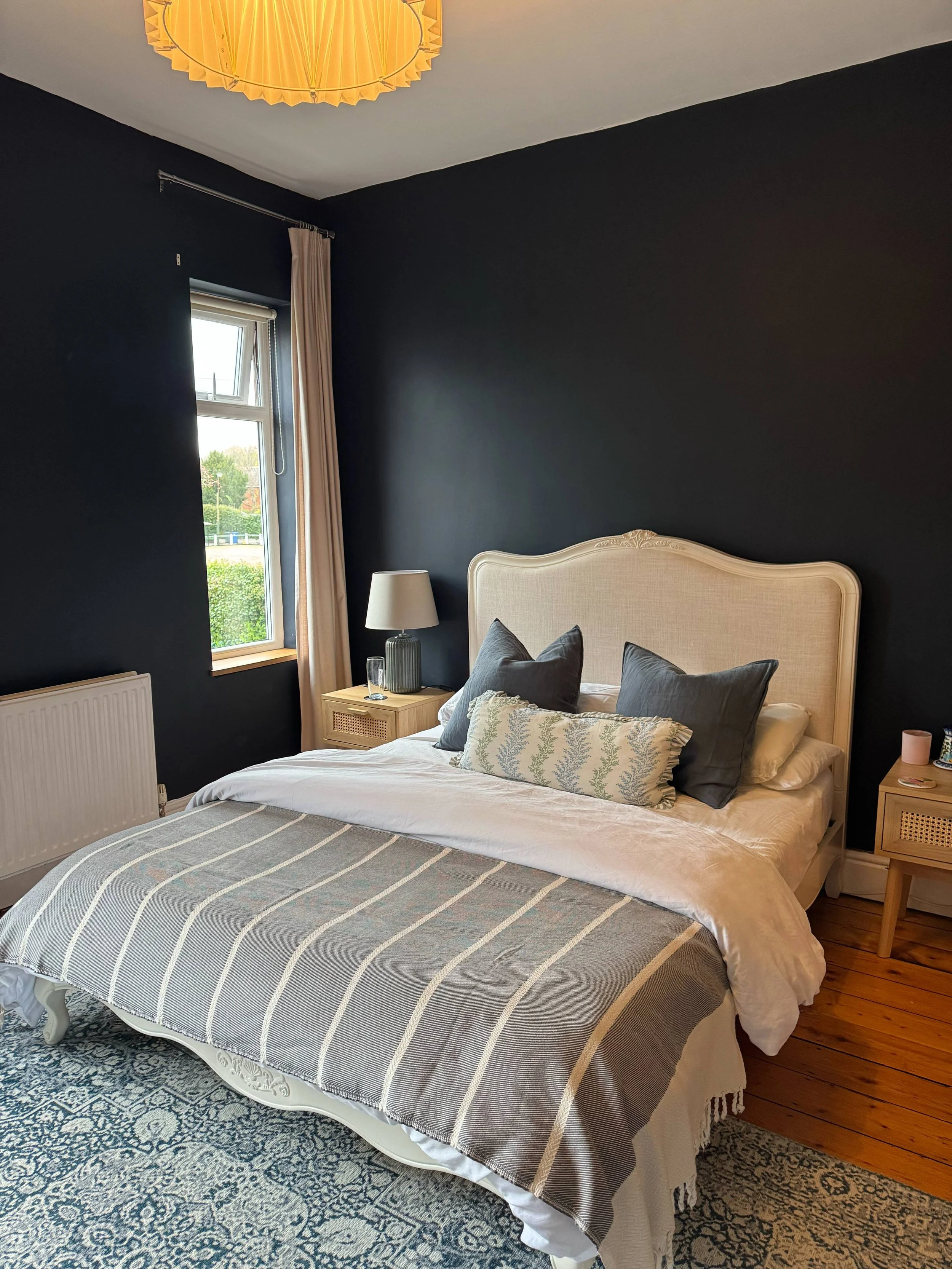 A bedroom with a bed, decorative pillows, nightstands, a lamp, a window with cream curtains, and a dark accent wall behind the bed.