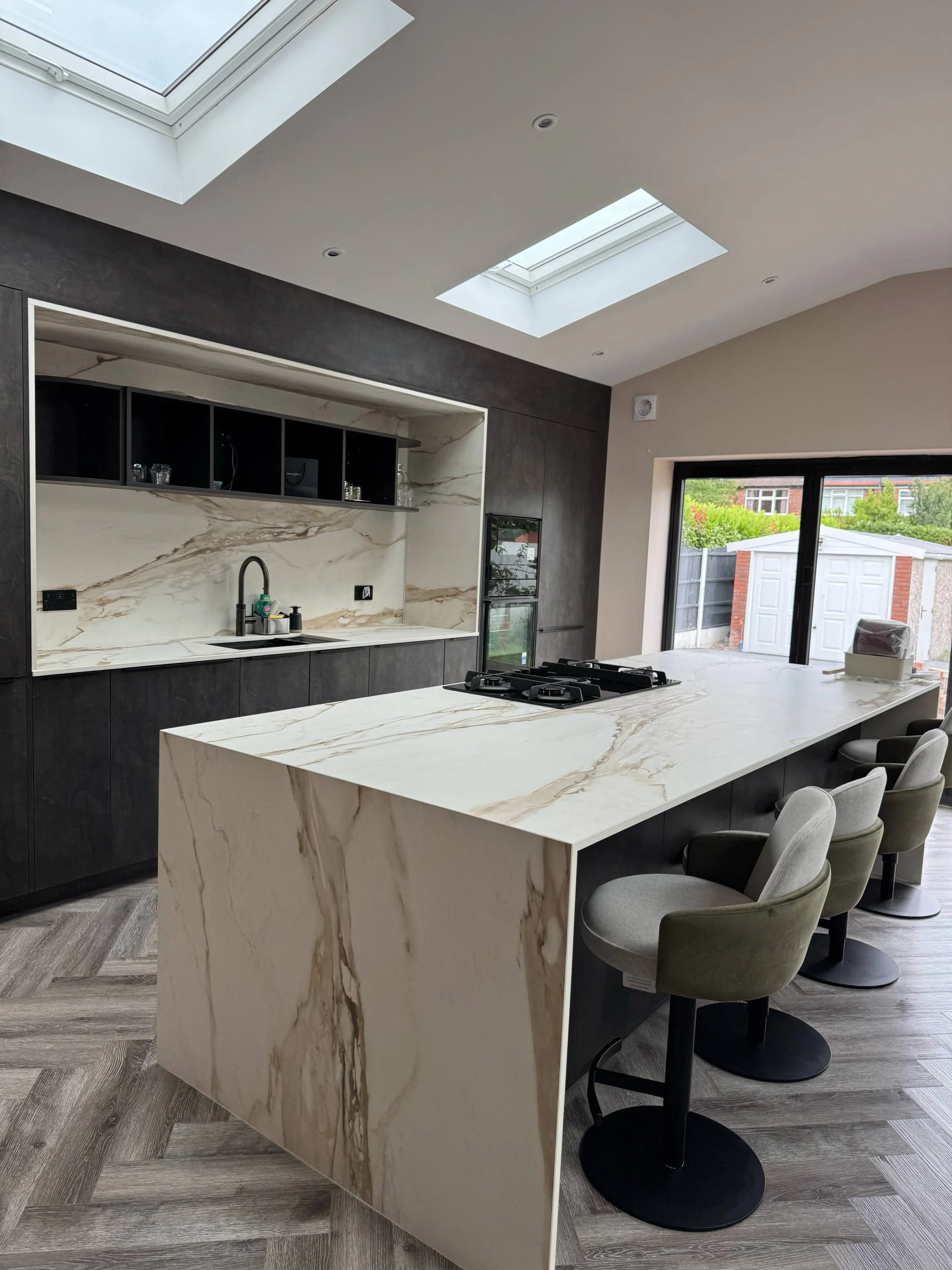 Modern kitchen with marble island, black cabinetry, large skylights, and sliding glass doors overlooking a backyard with a shed.