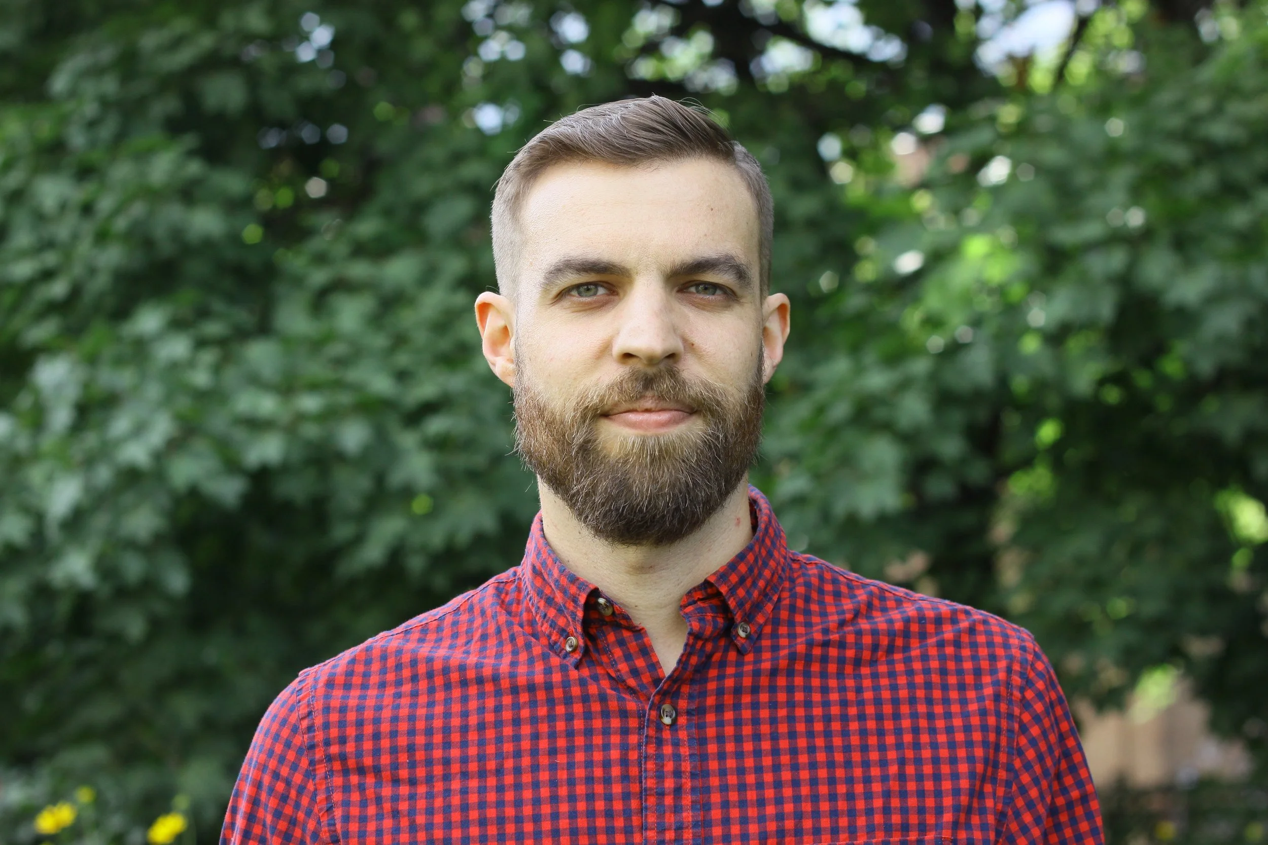 Jeune homme avec barbe portant une chemise à carreaux rouges et bleus, debout devant un fond de feuillage vert dense.