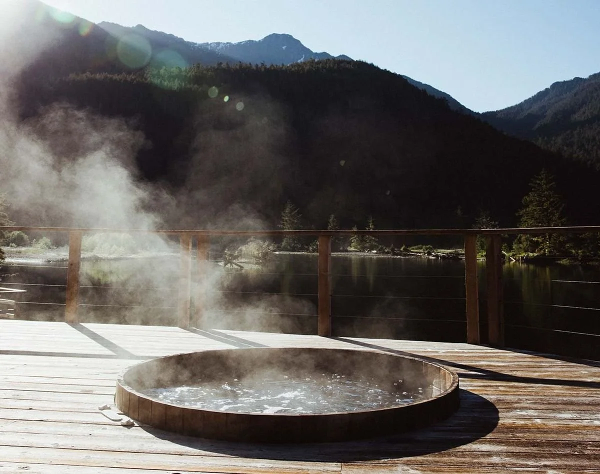 A steaming hot tub on a wooden deck overlooking a mountain lake with mist and trees.