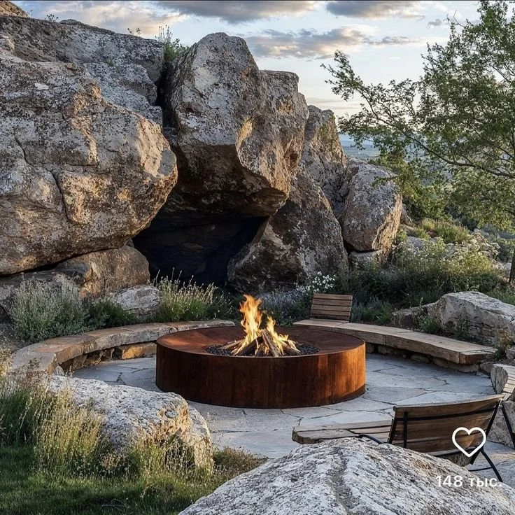A cozy outdoor seating area with a circular fire pit, wooden benches, and a chair surrounded by rocks and greenery.