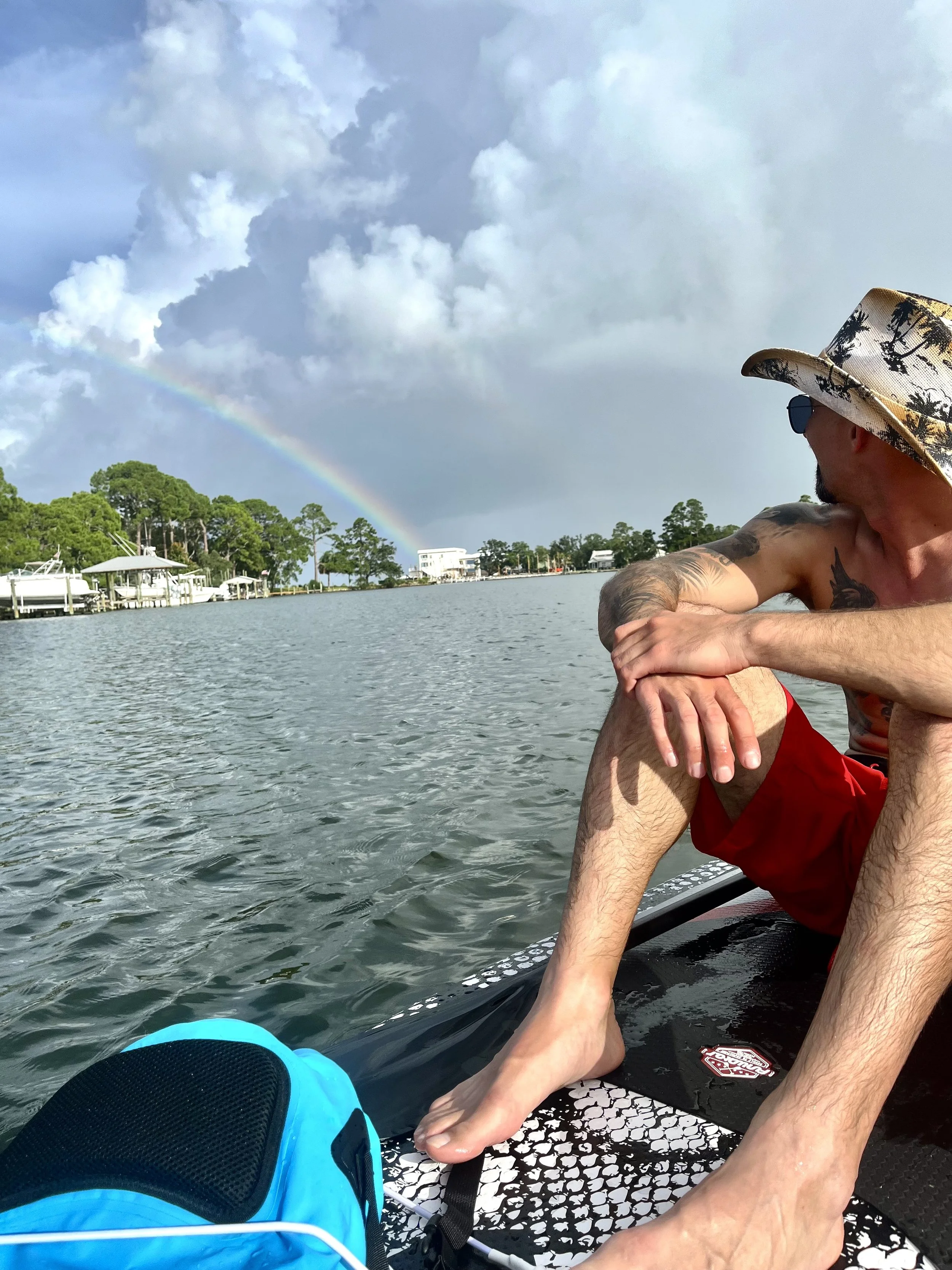 A man wearing a tropical hat and sunglasses sitting on a paddleboard on a lake, with a person’s hand touching his foot. In the background, there are boats, a dock, trees, and a rainbow in the cloudy sky.