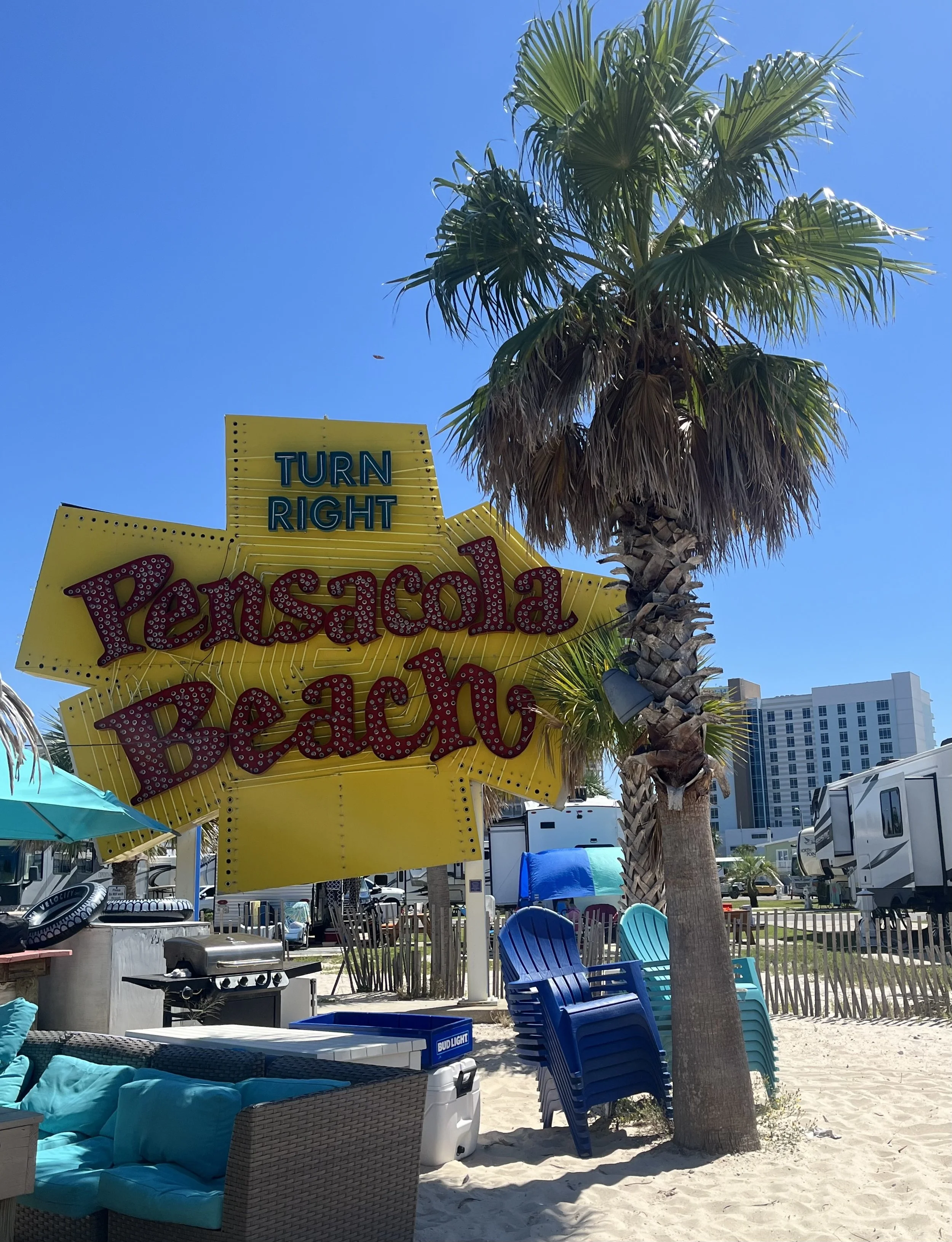 Beachside area with a tall palm tree, colorful chairs, a sign that says 'Turn Right Pensacola Beach,' and blue sky overhead.