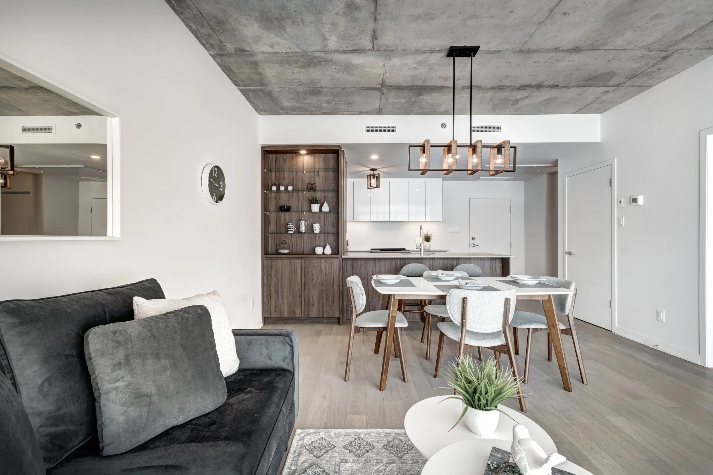 Modern living area with a gray couch, white pillows, and a white coffee table with decorative items, adjacent to a dining area with a wooden table and six chairs, with a wooden and glass shelving unit and a concrete ceiling.