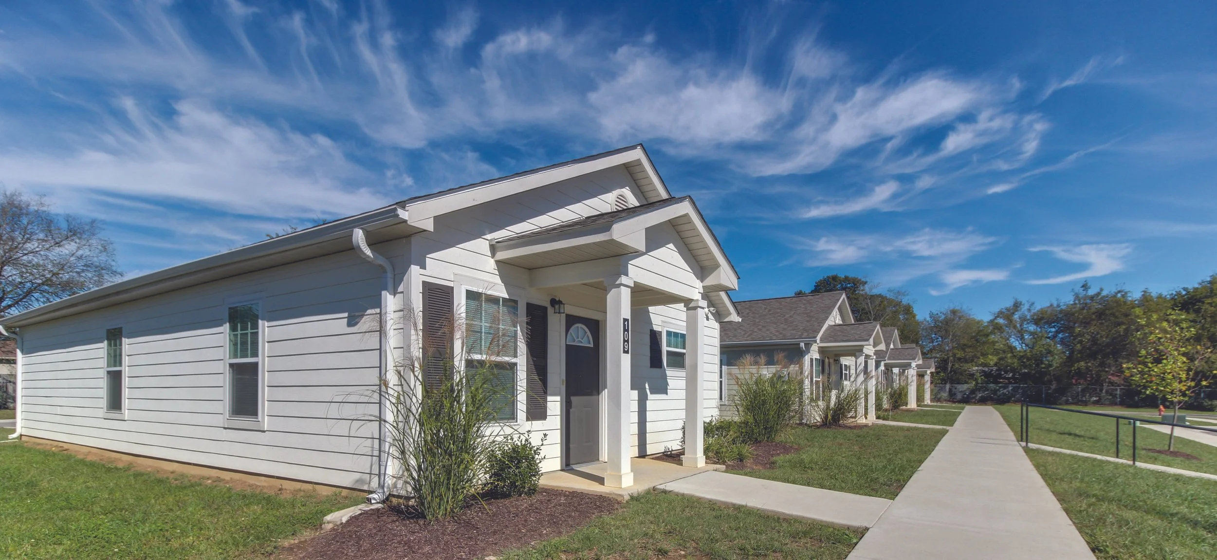 White residential building with front porch, black shutters, and lawn under a partly cloudy blue sky.