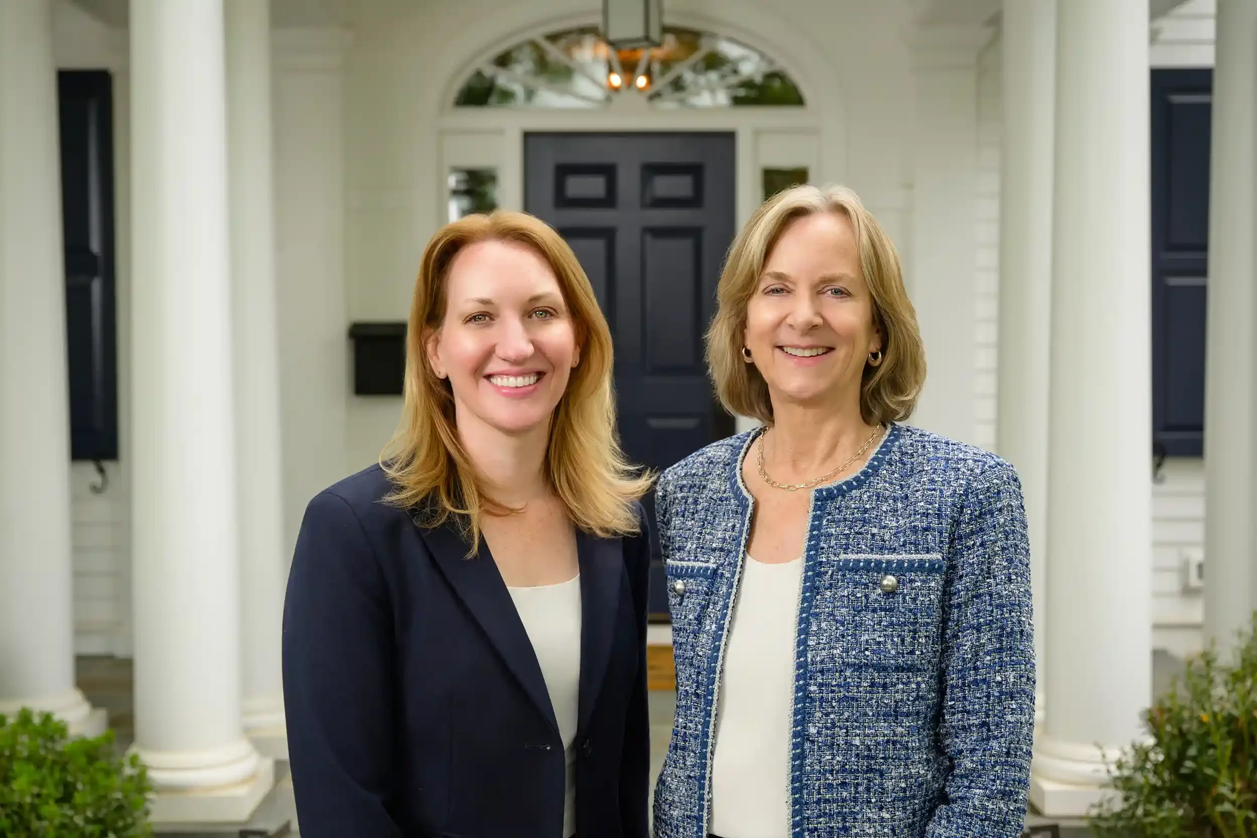 Allison Rivera and Kathy Rehill standing outside a house with white columns and a dark front door, smiling at the camera