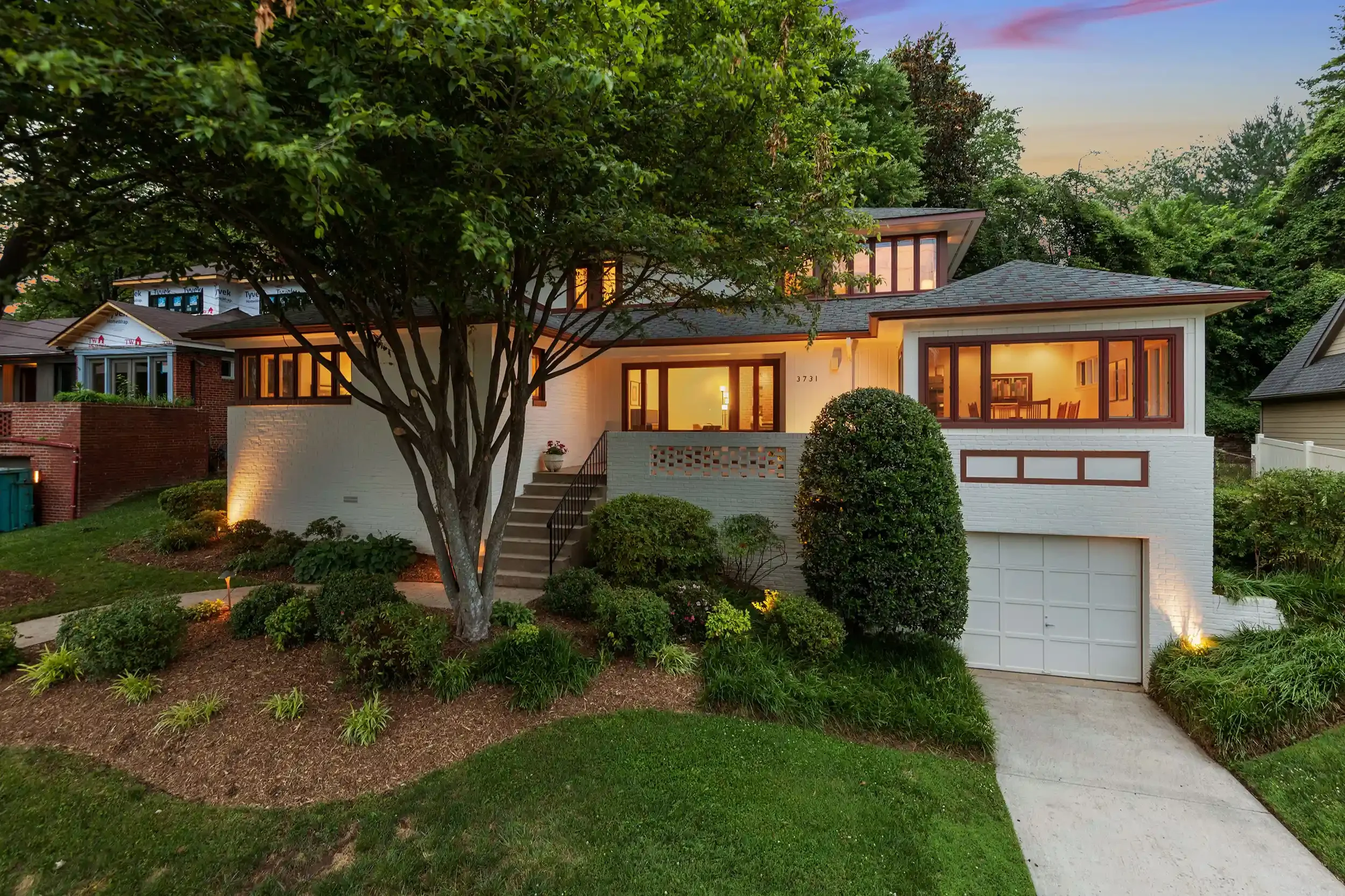 Front view of a modern house with large windows, a white exterior, and a landscaped yard with bushes and a large tree, illuminated by warm lighting at dusk