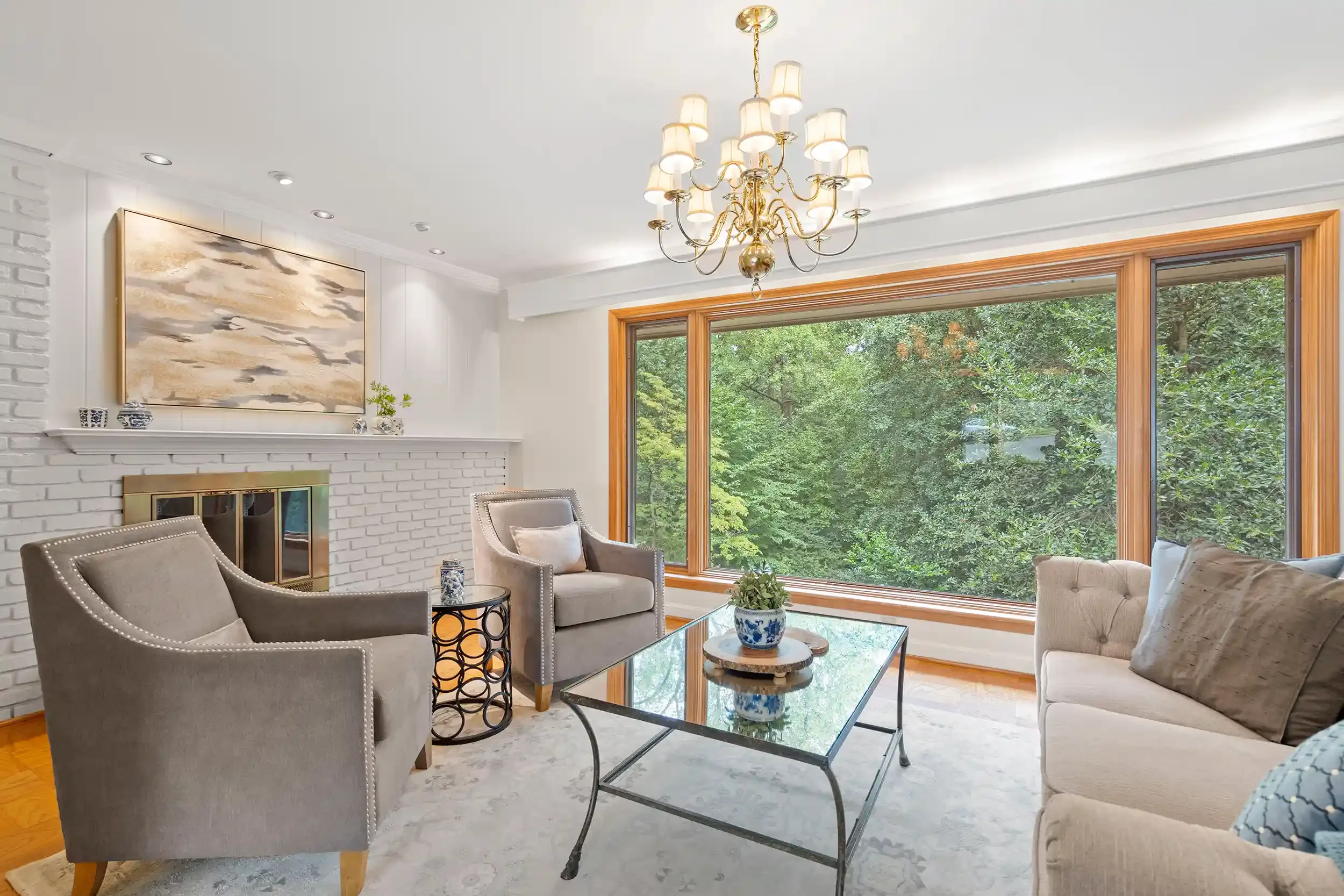 A living room with large windows showing trees outside, a white brick fireplace, neutral-colored furniture, a glass coffee table, and a gold chandelier with multiple lamps
