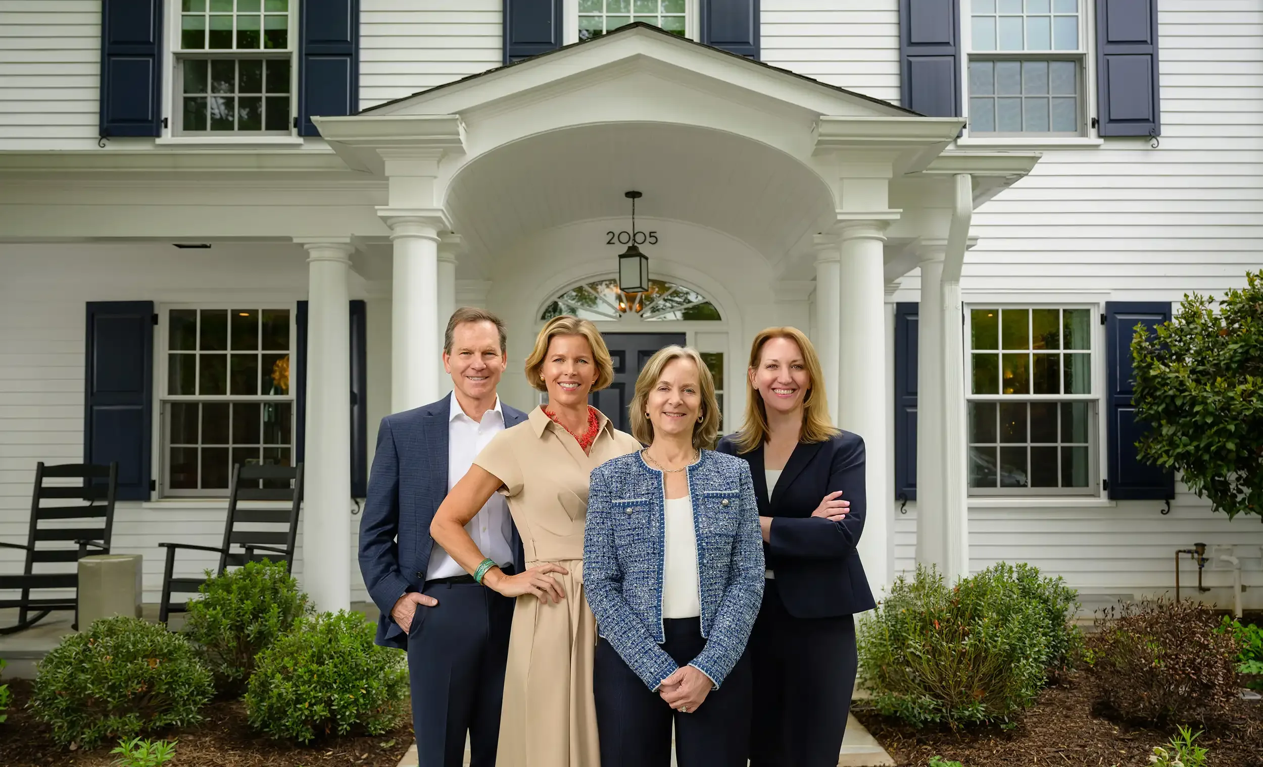The Beall & Rehill team standing in front of a white house with columns, windows, and blue shutters, smiling at the camera