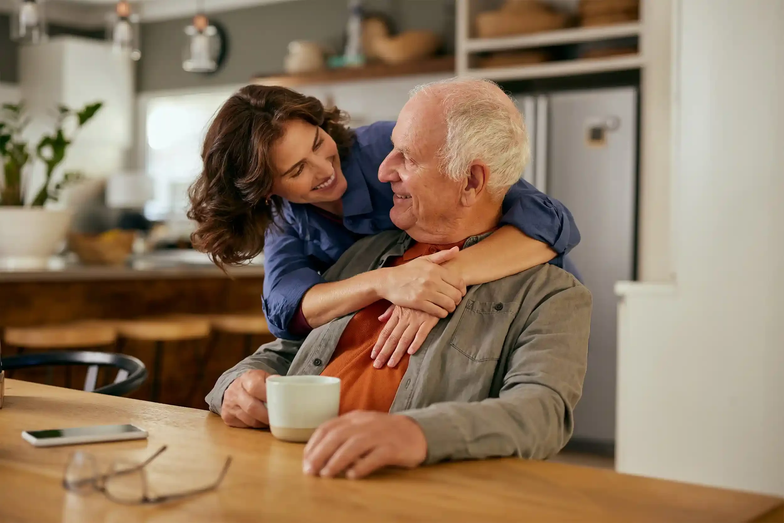 A daughter with brown curly hair hugging and smiling at her elderly father with white hair