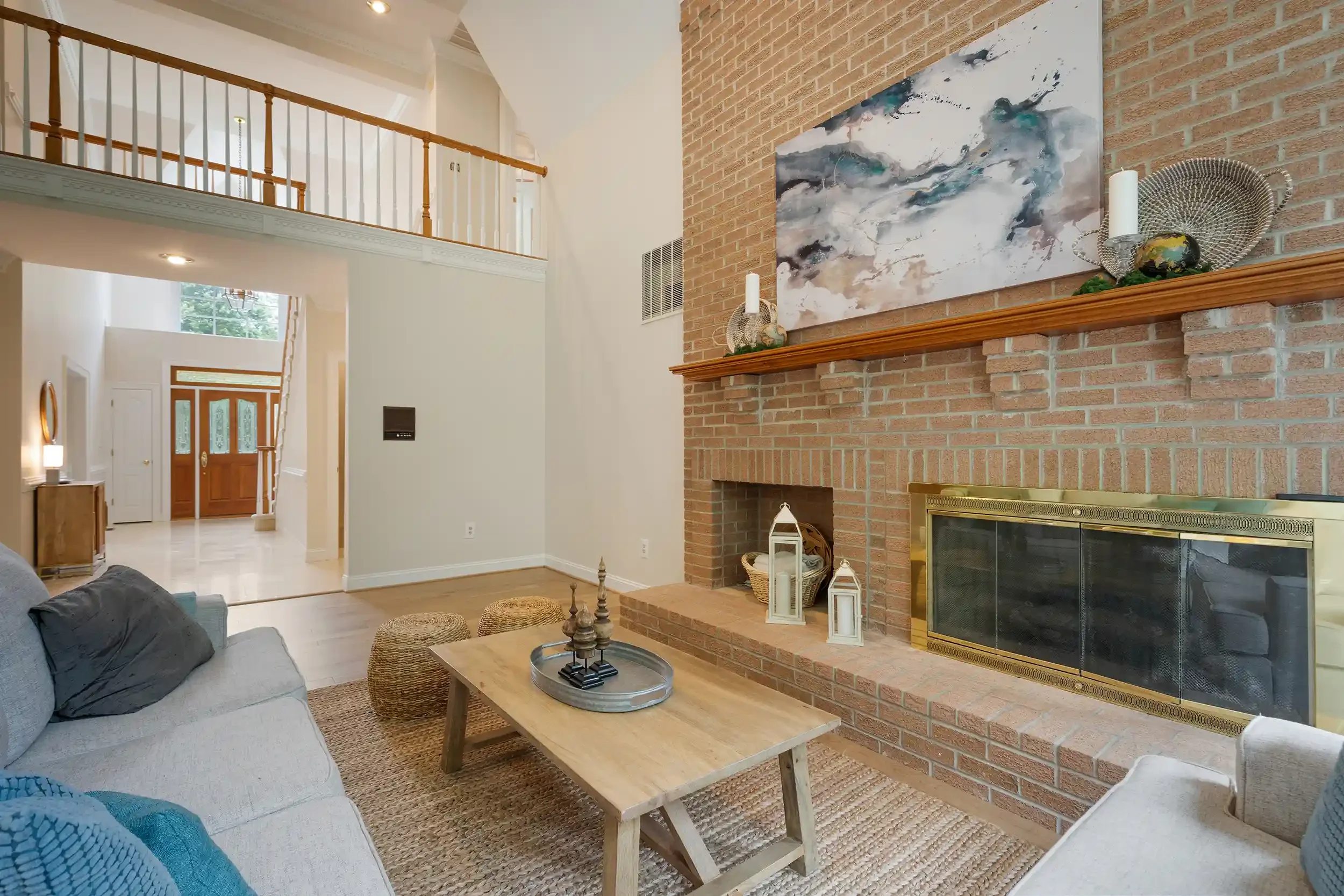 Living room with brick fireplace, abstract painting, wooden mantel, candles, wicker baskets, and a gray couch with blue pillows