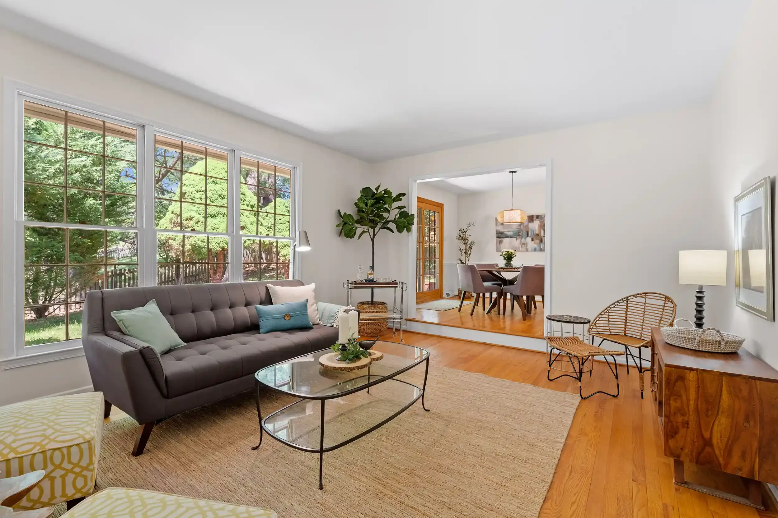 Bright living room with large windows showing green trees outside, featuring a gray tufted sofa with pillows, a glass coffee table, and a woven rug on hardwood floor, with a connected dining area visible in the background